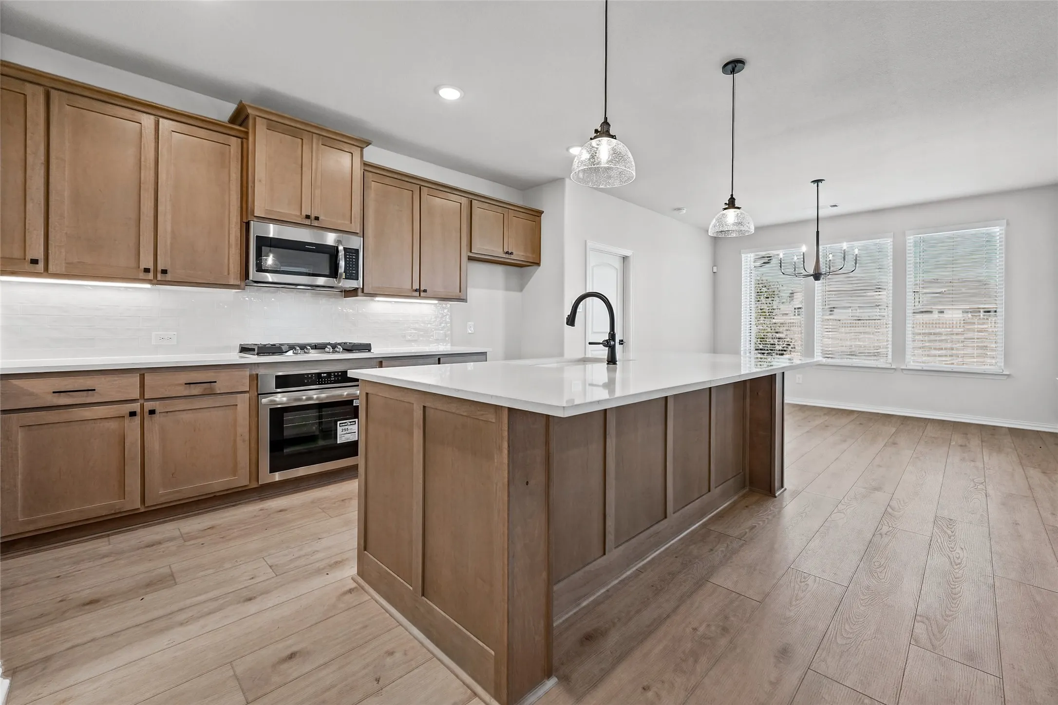 Wood-finish flooring throughout, kitchen featuring light wood cabinetry, stainless steel appliances, a white tile backsplash, and a central island with a solid surface countertop