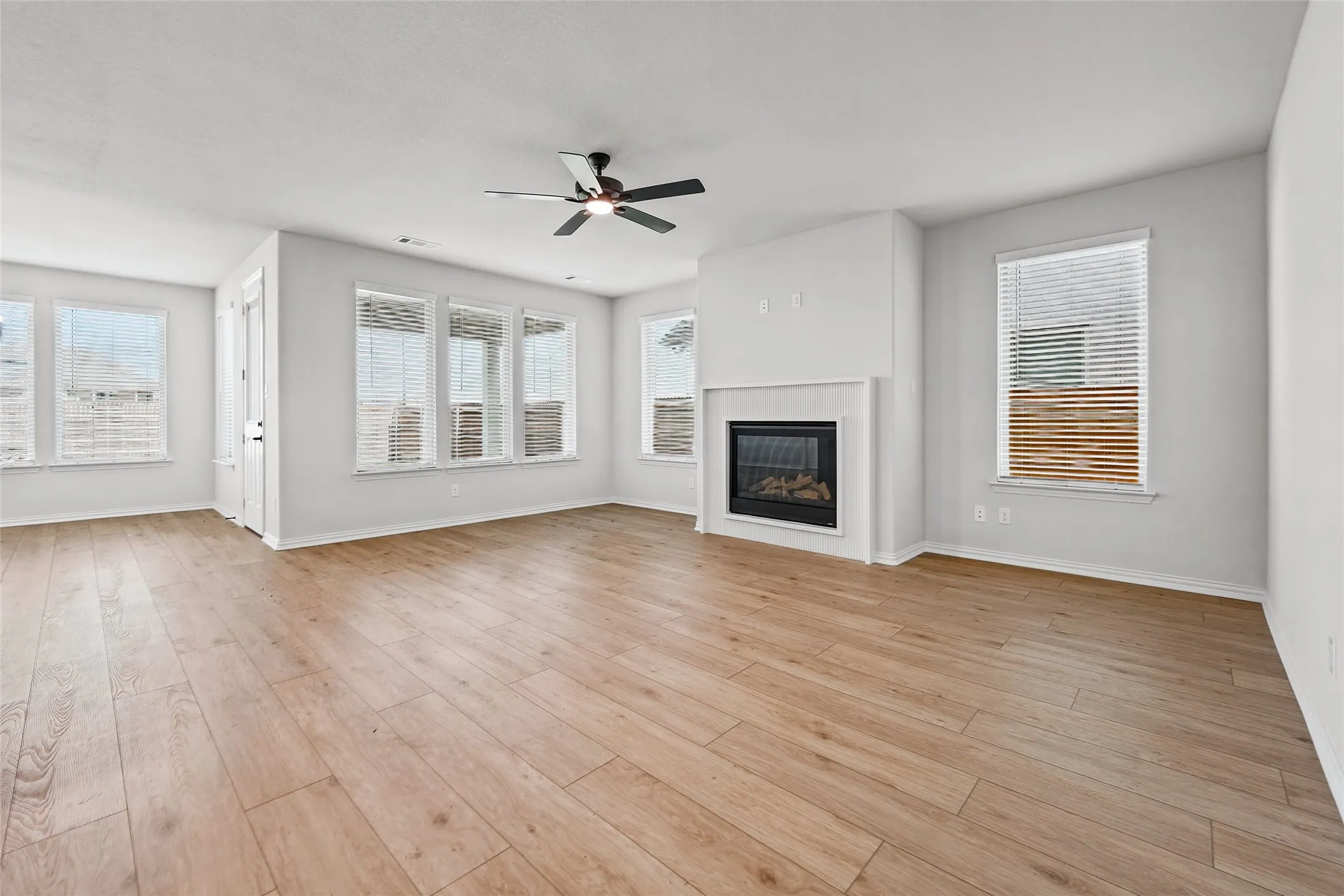 Spacious living area featuring wood-finish flooring, a built-in fireplace with a mantel, multiple windows with blinds, and a ceiling fan