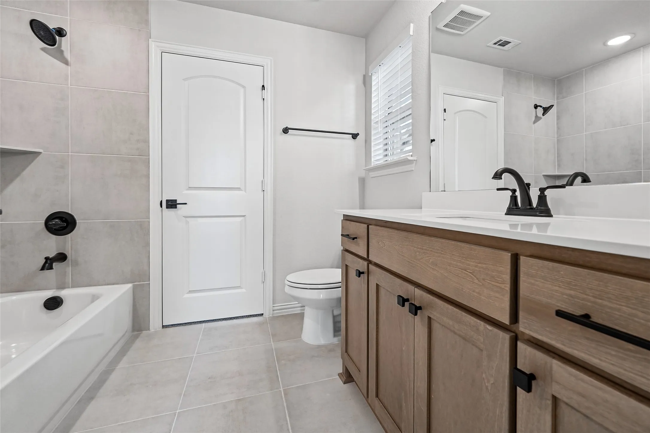 White bathtub with matte black fixtures, light-toned wall tiling, wood-finish vanity with matte black hardware, white countertop, and large vanity mirror