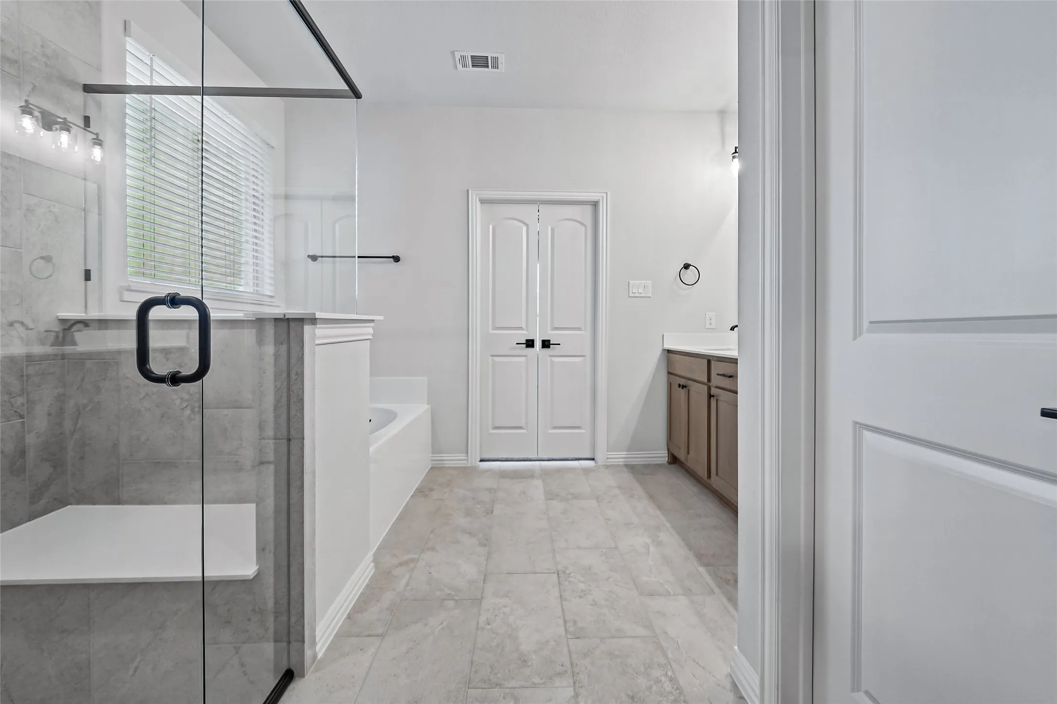 Spacious bathroom with tiled flooring, a glass-enclosed shower featuring a built-in bench, a separate soaking tub, and a double vanity with a light wood-finish cabinet