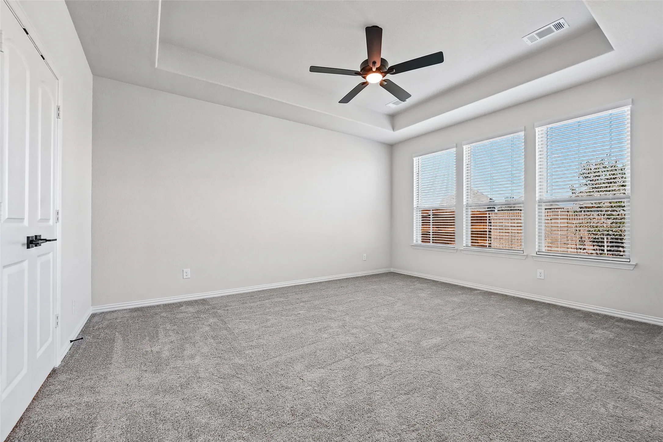 Recessed ceiling with an attached ceiling fan, three paneled windows with horizontal blinds, and grey carpet flooring