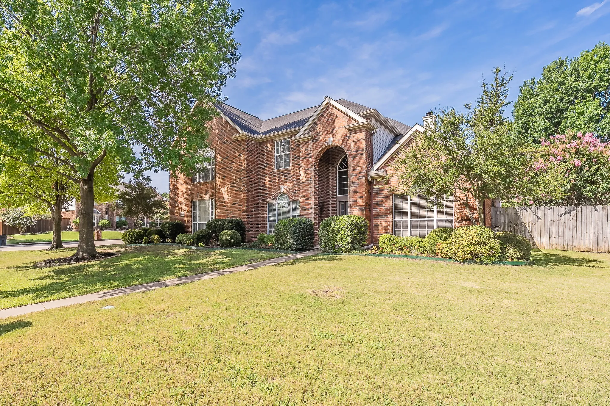 Brick exterior residence featuring an arched entryway, multiple windows with grilles, established landscaping, and a partial privacy fence