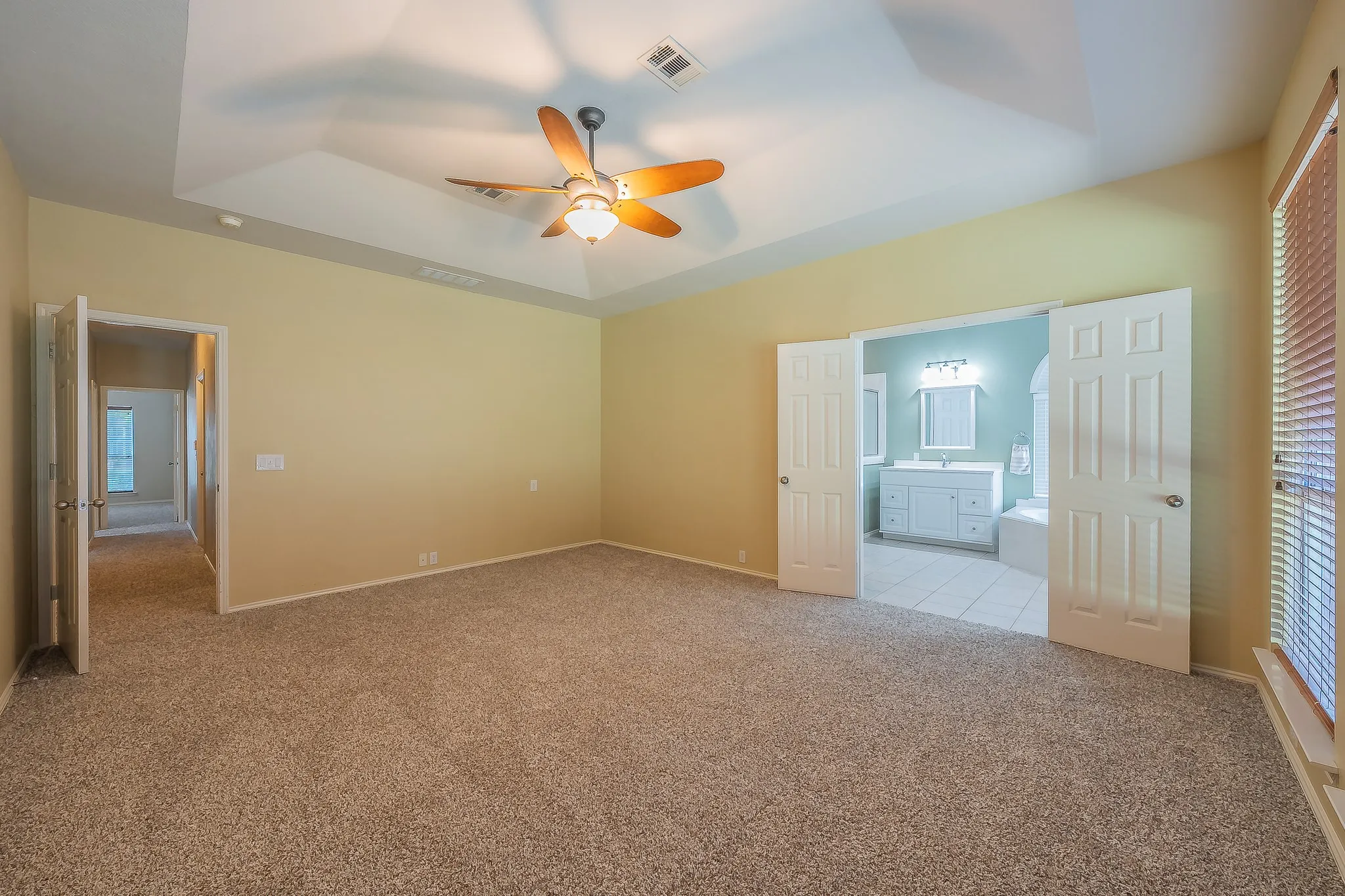 Spacious room featuring neutral carpeting, a tray ceiling with a ceiling fan, and a large window with blinds