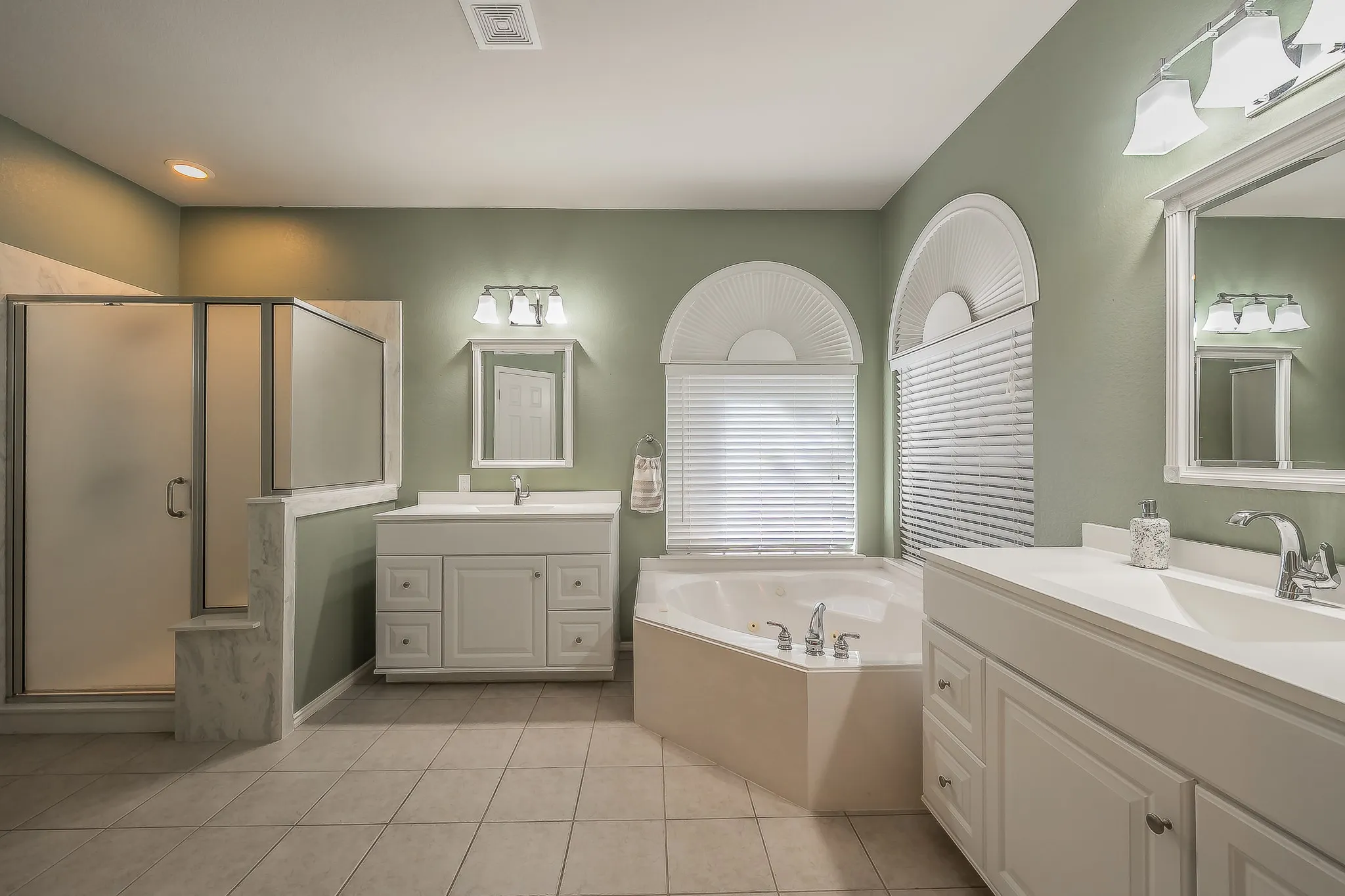 Spacious bathroom featuring an oversized corner soaking tub, a tiled walk-in shower with frosted glass enclosure, and dual vanity sinks with white cabinetry