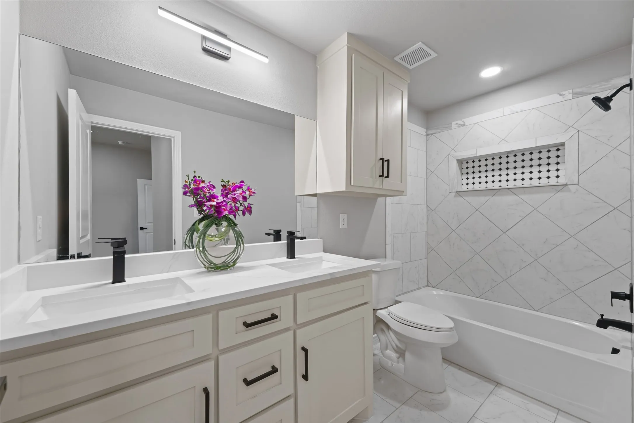 Bathroom featuring a double vanity with matte black fixtures, an expansive mirror with integrated lighting, a white bathtub, and a tiled shower surround with a decorative niche