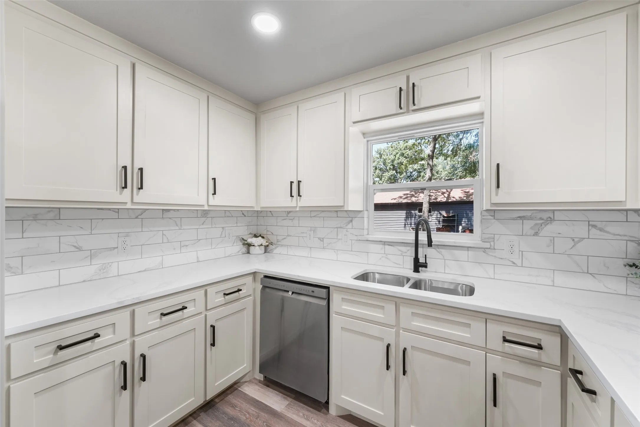 Modern kitchen featuring white shaker cabinetry, an undermount stainless steel double sink, a black gooseneck faucet, light-toned countertops, and a white subway tile backsplash with veining