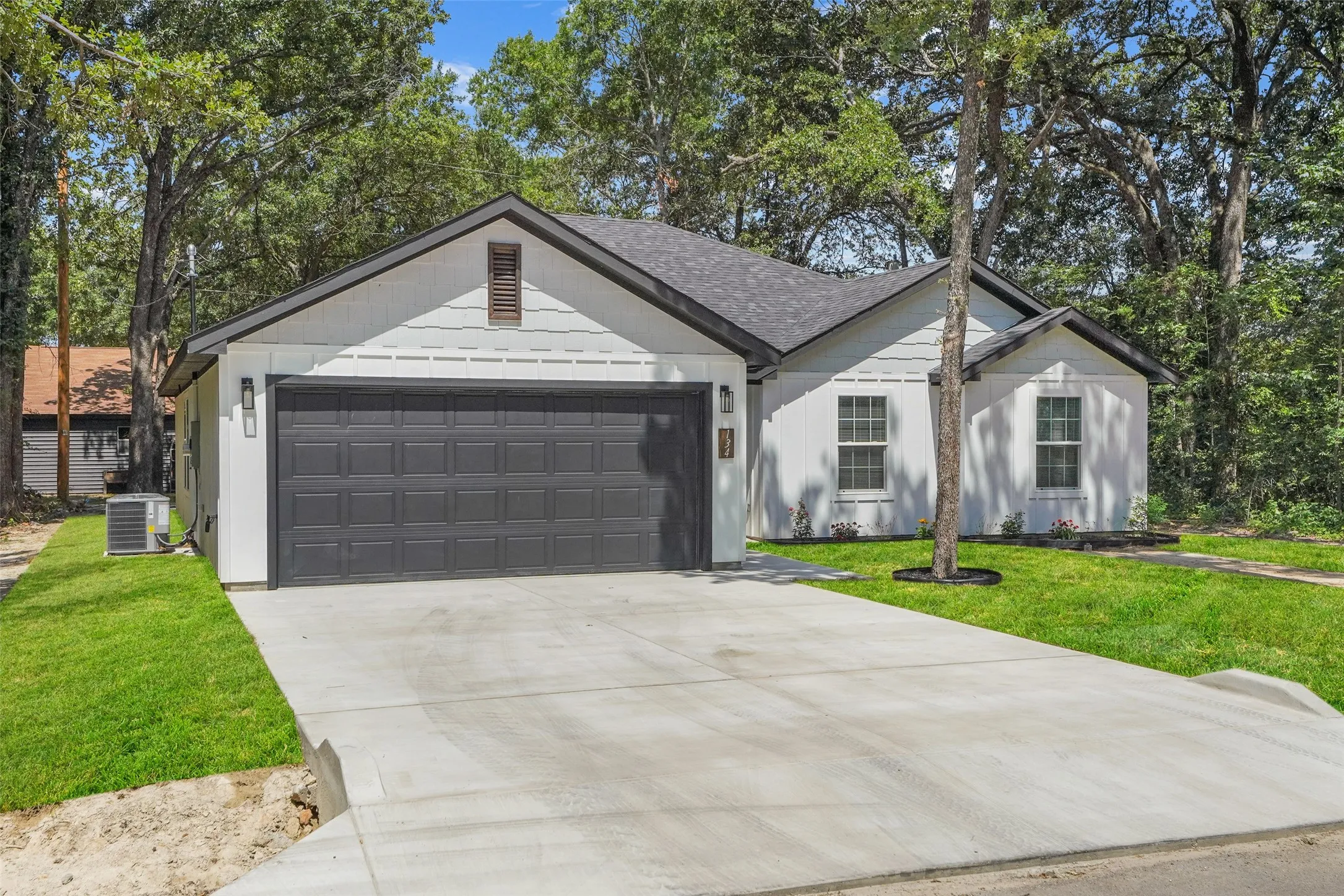 Contemporary single-story residence featuring a dark gray two-car garage door, white horizontal siding, a dark gray shingled roof, a concrete driveway, and established trees