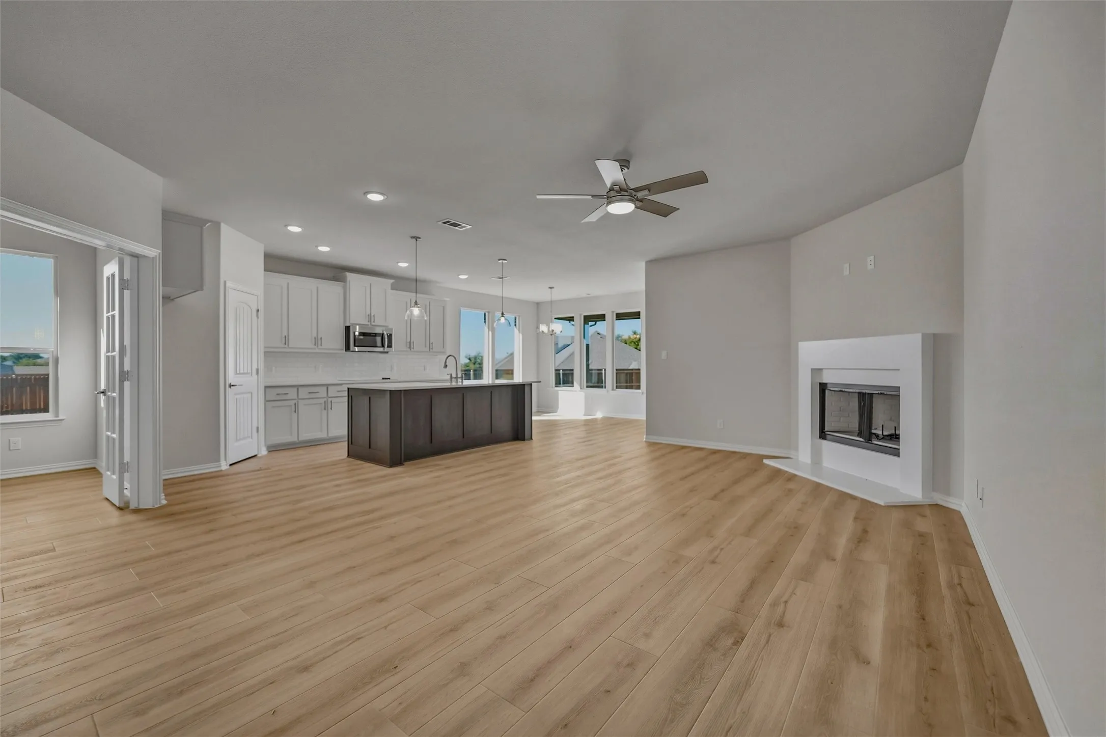 Open-concept living space featuring wood-finish flooring, a white-mantel fireplace, a ceiling fan, a kitchen island with pendant lighting, and white cabinetry