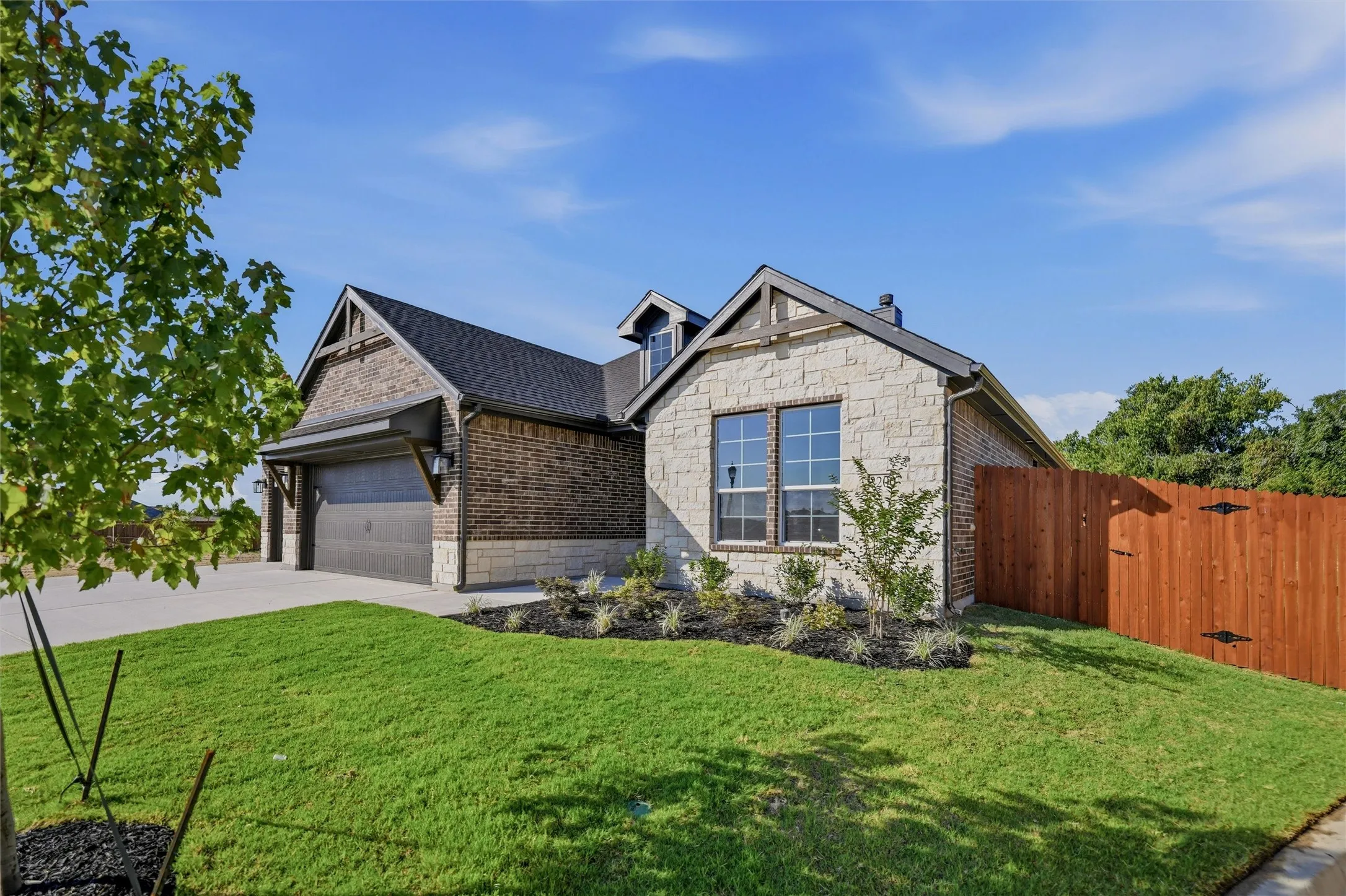 Stone and brick exterior with dark trim accents, featuring a two-car garage, established landscaping, and a wood privacy fence