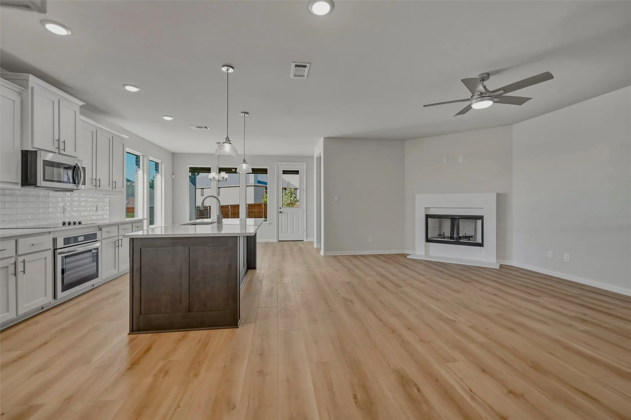 Open concept living space featuring wood-finish flooring, a kitchen island with pendant lighting, white cabinetry, a built-in oven, and a fireplace with a mantel