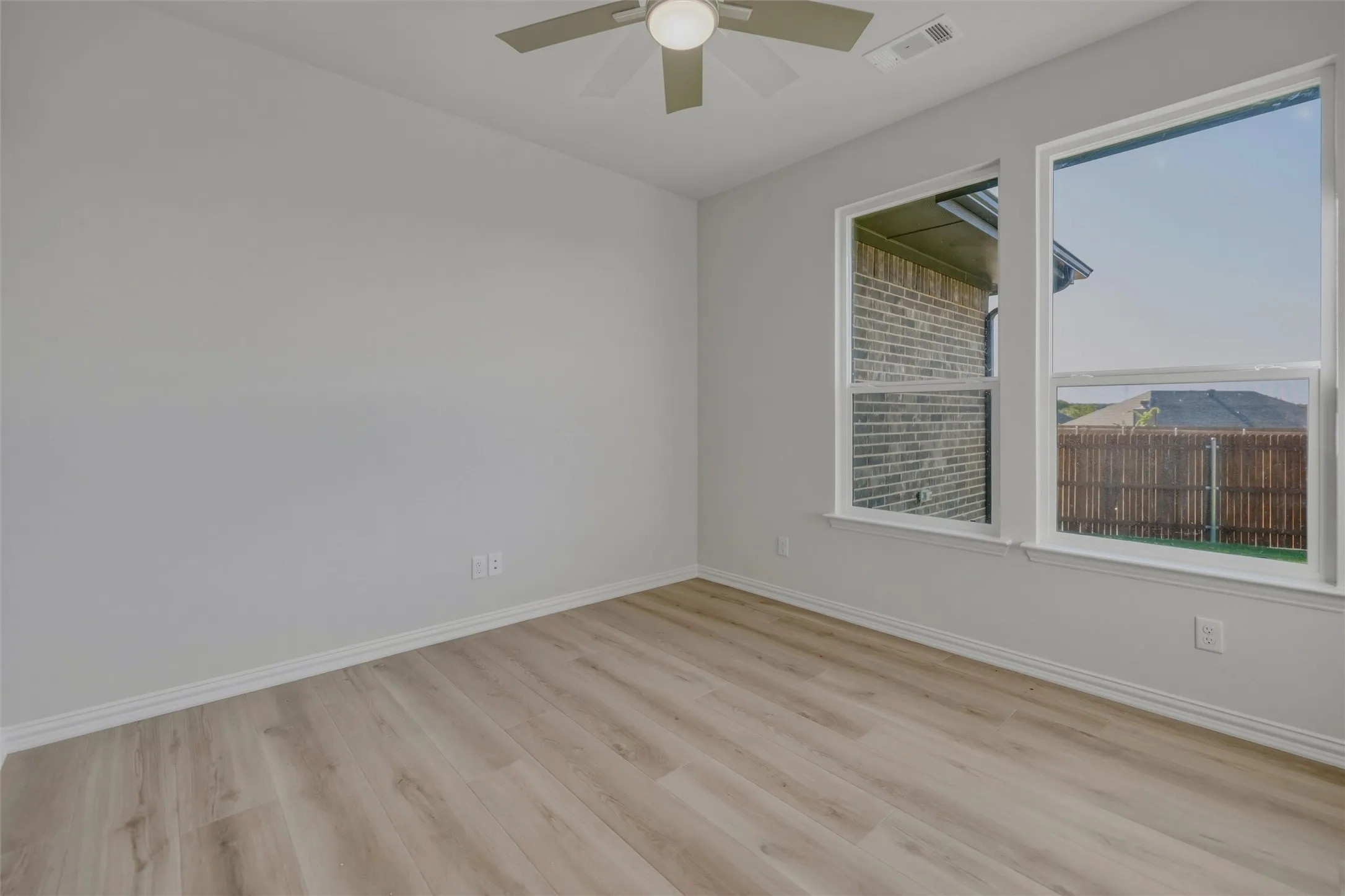 Light wood-finish flooring, two large windows, white trim, and a ceiling fan with integrated lighting
