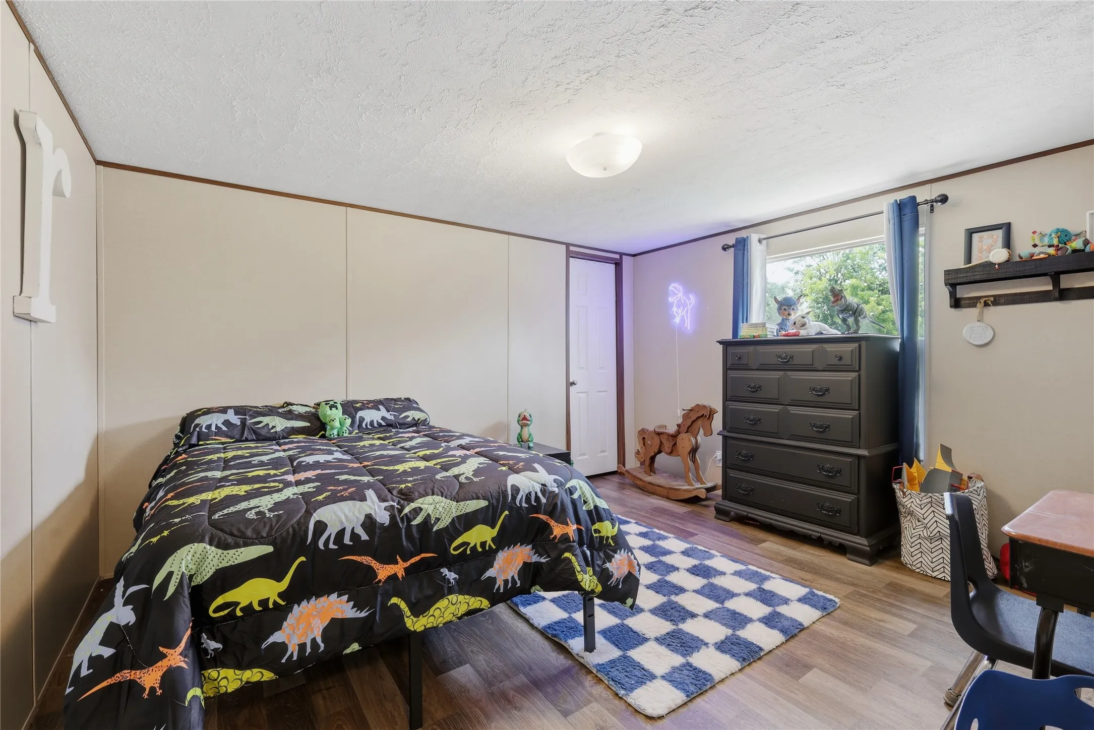Room featuring a neutral wall paneling, a single window with blue curtains, and wood-finish flooring