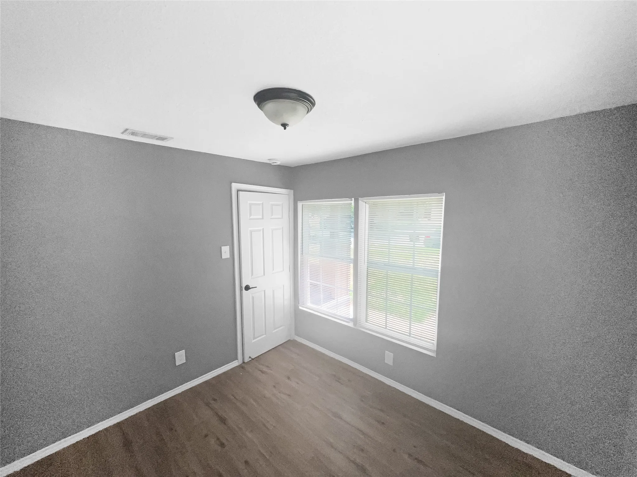 Room with wood-finish flooring, two large windows with blinds, a white paneled door with black hardware, recessed ceiling light, and gray walls