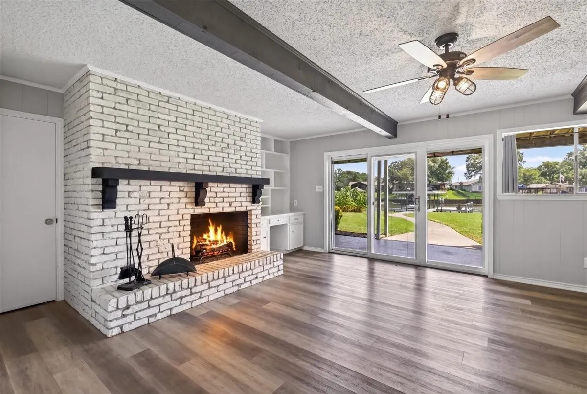 Expansive downstairs walk-out basement game room living area featuring wood-finish flooring, a white brick fireplace with a black mantel, built-in shelving, and direct outdoor access via sliding glass doors to the lake side yard.  Downstairs also has a large cedar lined storage closet, bedroom with ensuite bathroom and walk-in closet