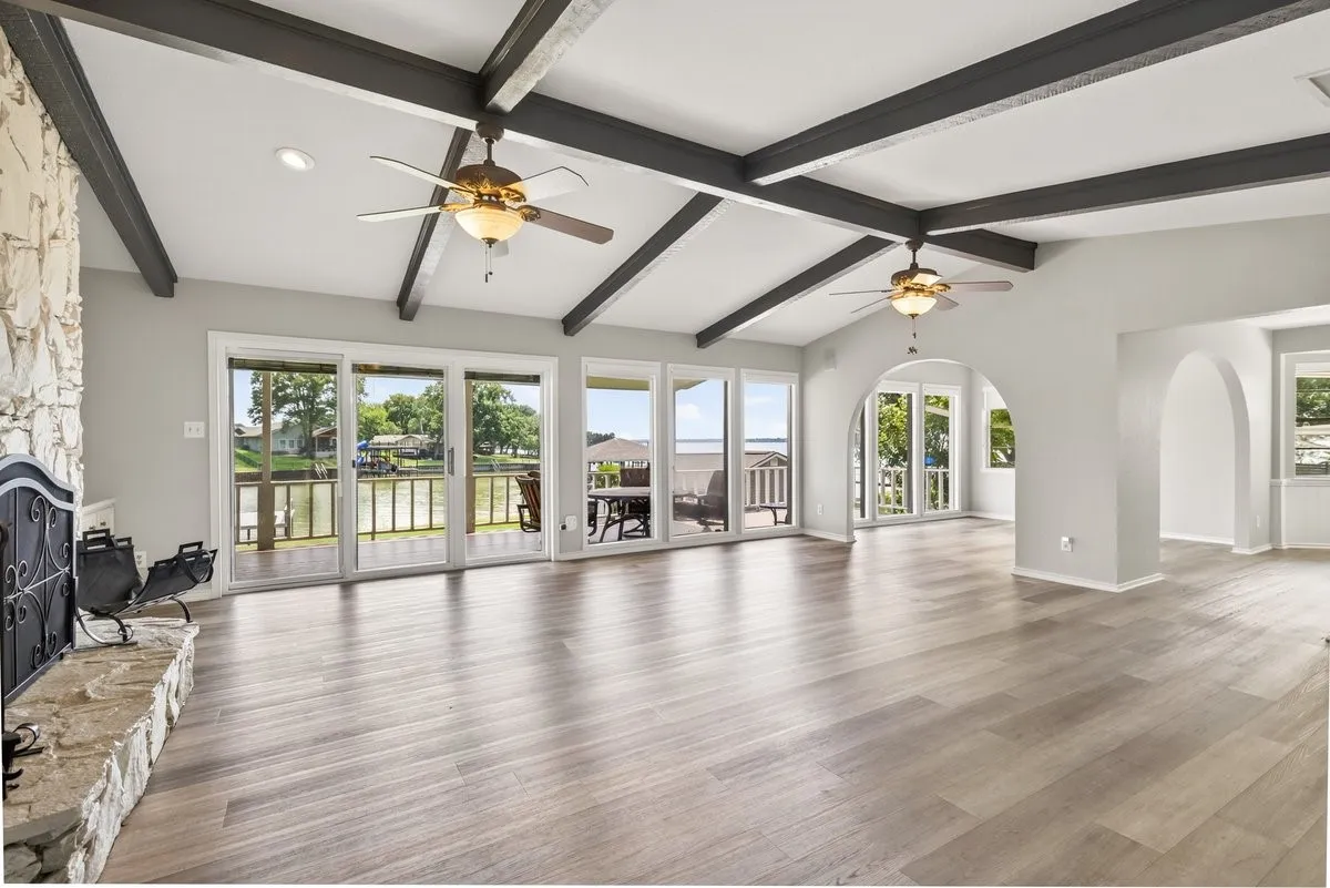 Spacious living area with expansive lake view through floor to ceiling windows while featuring multi-tone wood-finish flooring and a vaulted ceiling with exposed dark wood beams, open to the dining room and kitchen entry