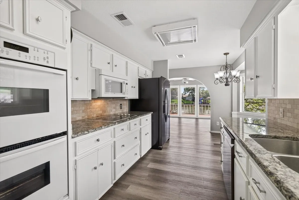 Galley kitchen view facing toward lakeside dining room, featuring white cabinetry, granite countertops, tile backsplash, stainless steel double sink, double ovens, cooktop, and wood-finish flooring