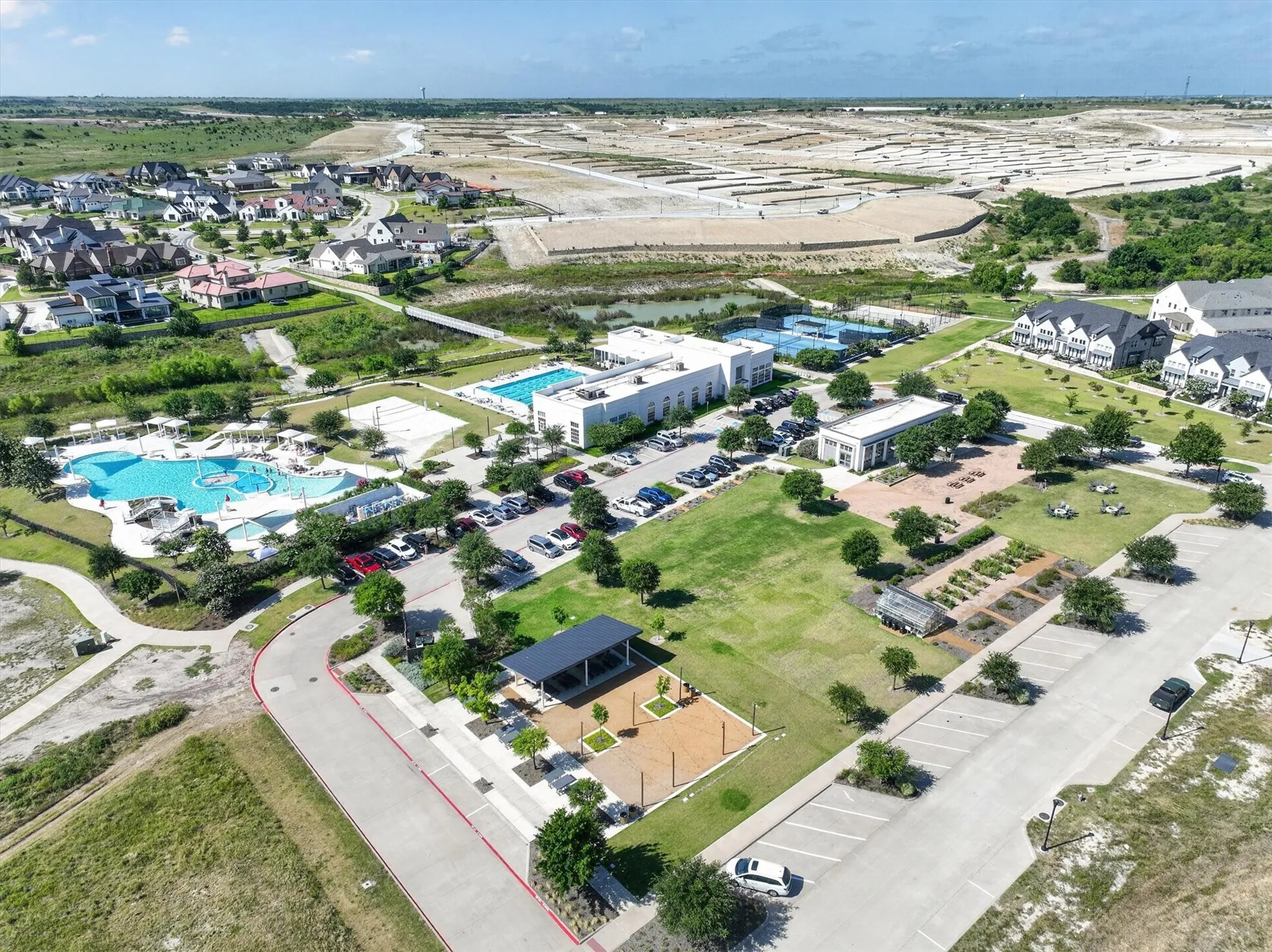 Aerial perspective showcasing a comprehensive community featuring multiple swimming pools, tennis courts, a central green space, and structured parking areas