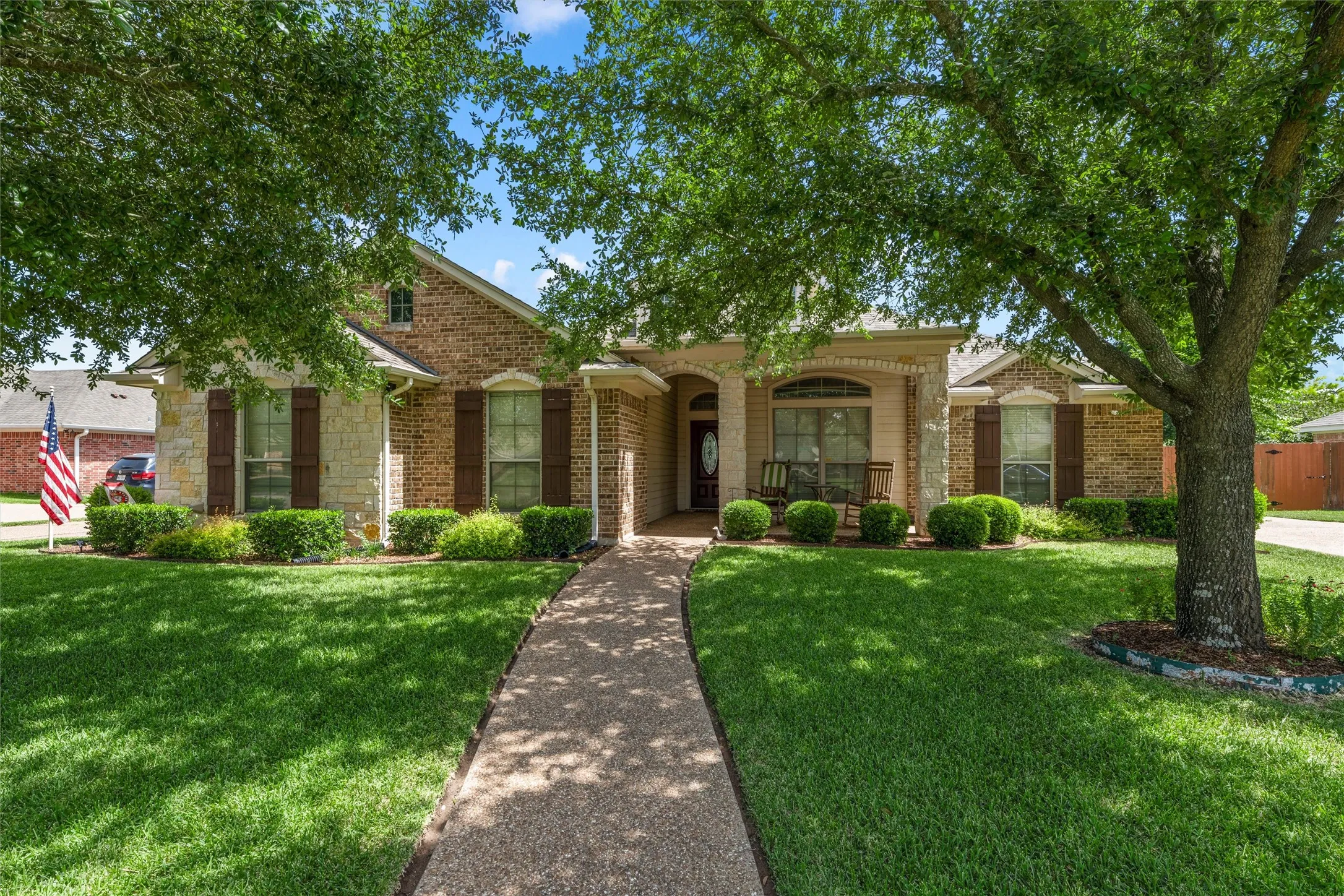 Brick and stone facade residence featuring a covered front porch, mature trees, manicured lawn, and a patterned concrete walkway