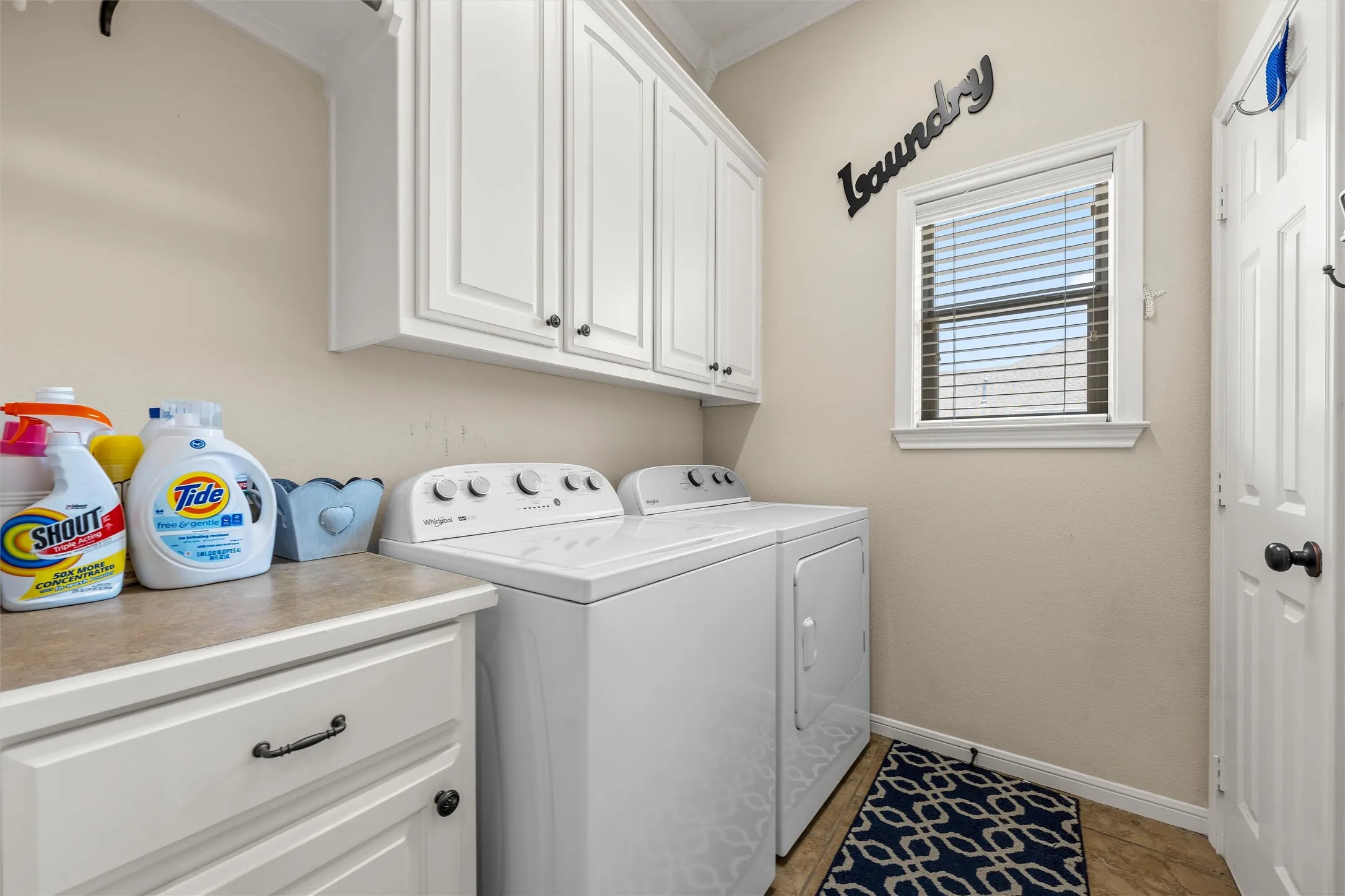Dedicated laundry space featuring upper and lower cabinetry with black hardware, a window with horizontal blinds, and a tiled floor