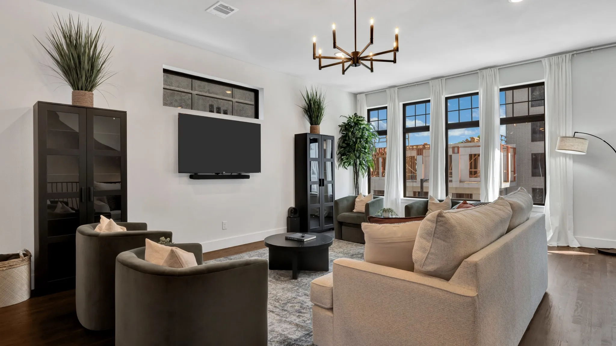 Spacious living area featuring dark wood-finish flooring, a modern chandelier, and multiple tall windows with white drapes