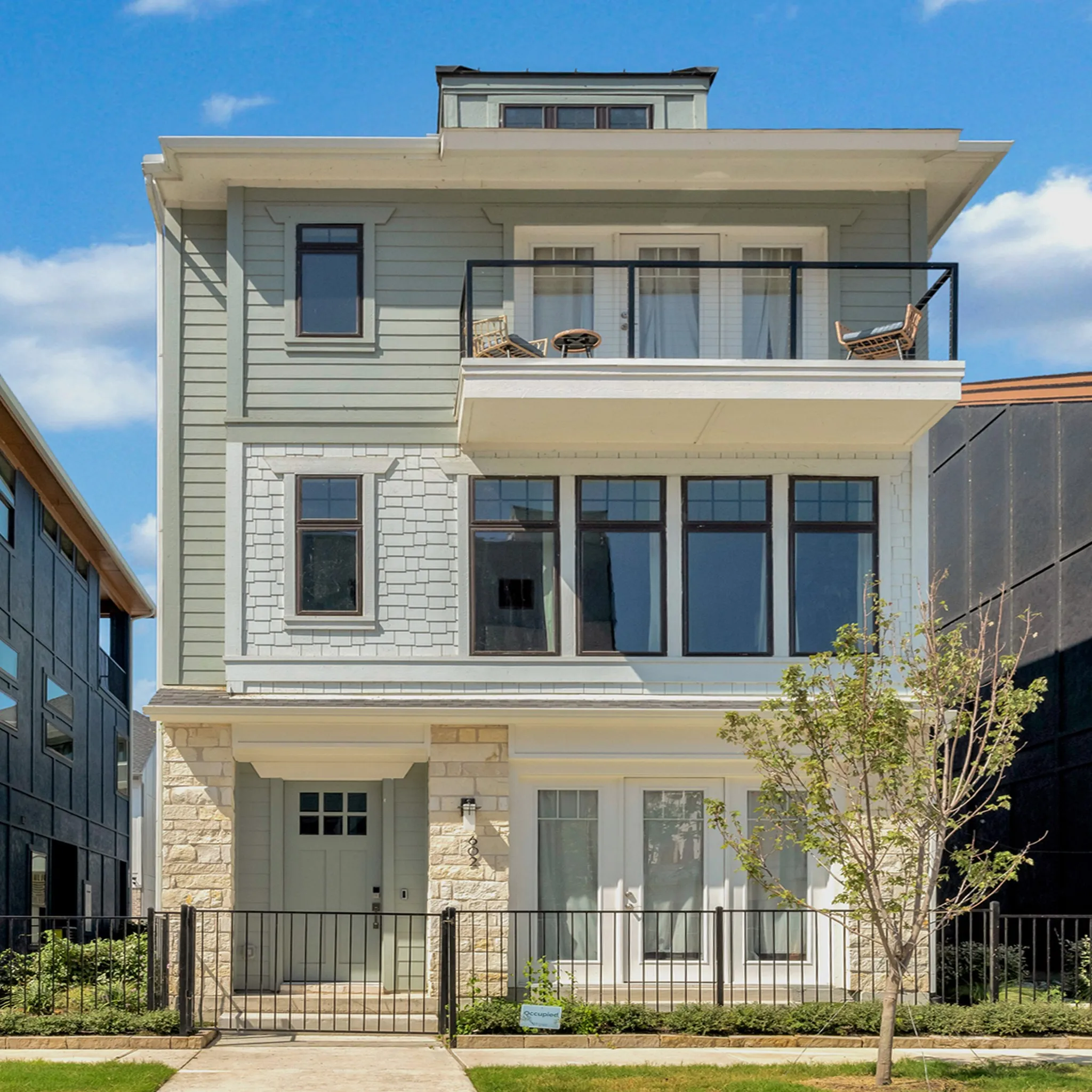 Contemporary multi-story facade featuring light green siding, white shingle accents, stone veneer, a second-story balcony with metal railing, and a black metal fence enclosing the front yard