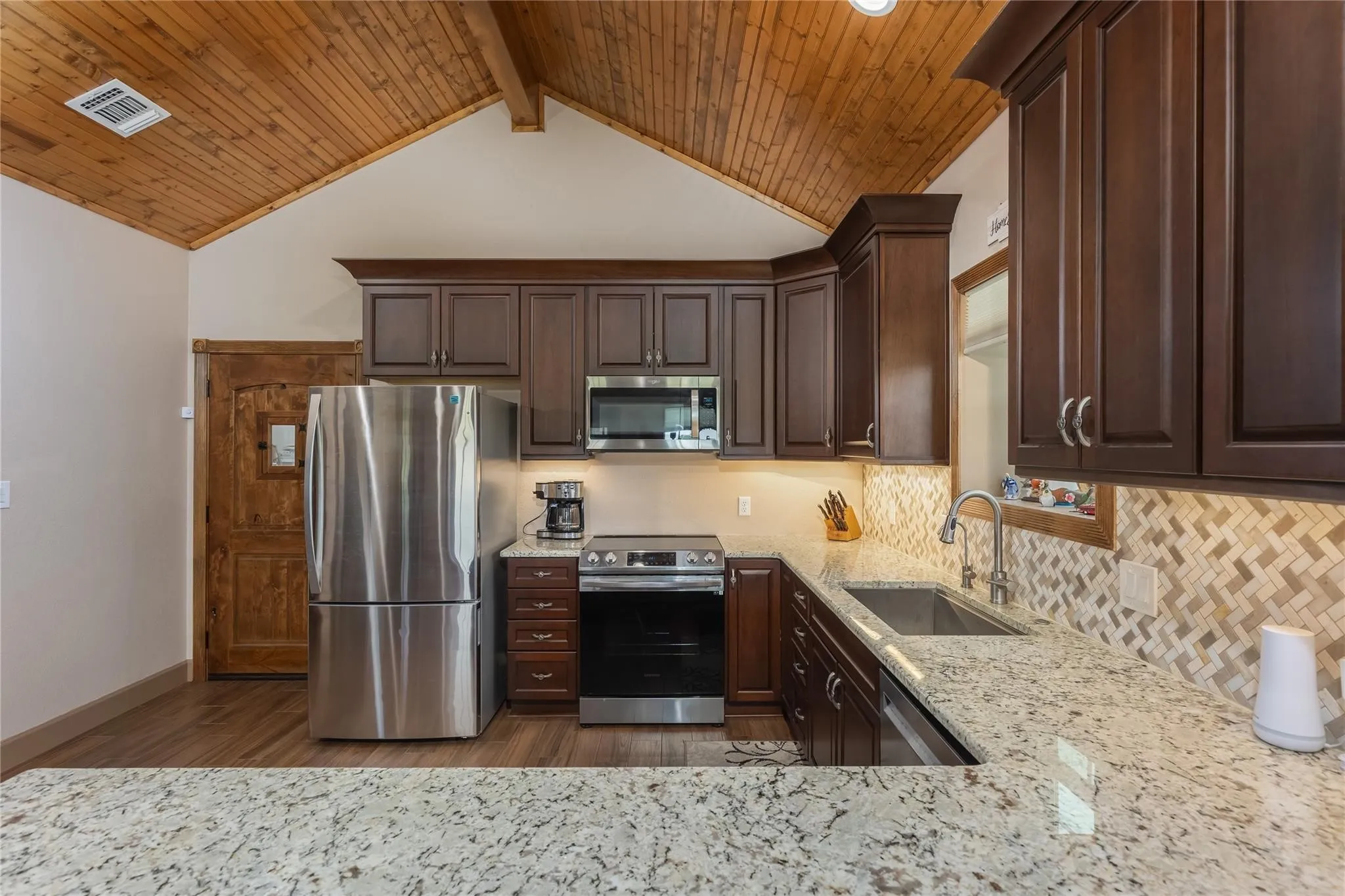 Kitchen featuring a vaulted wood-paneled ceiling, dark wood cabinetry, stainless steel appliances, light granite countertops, and a patterned tile backsplash