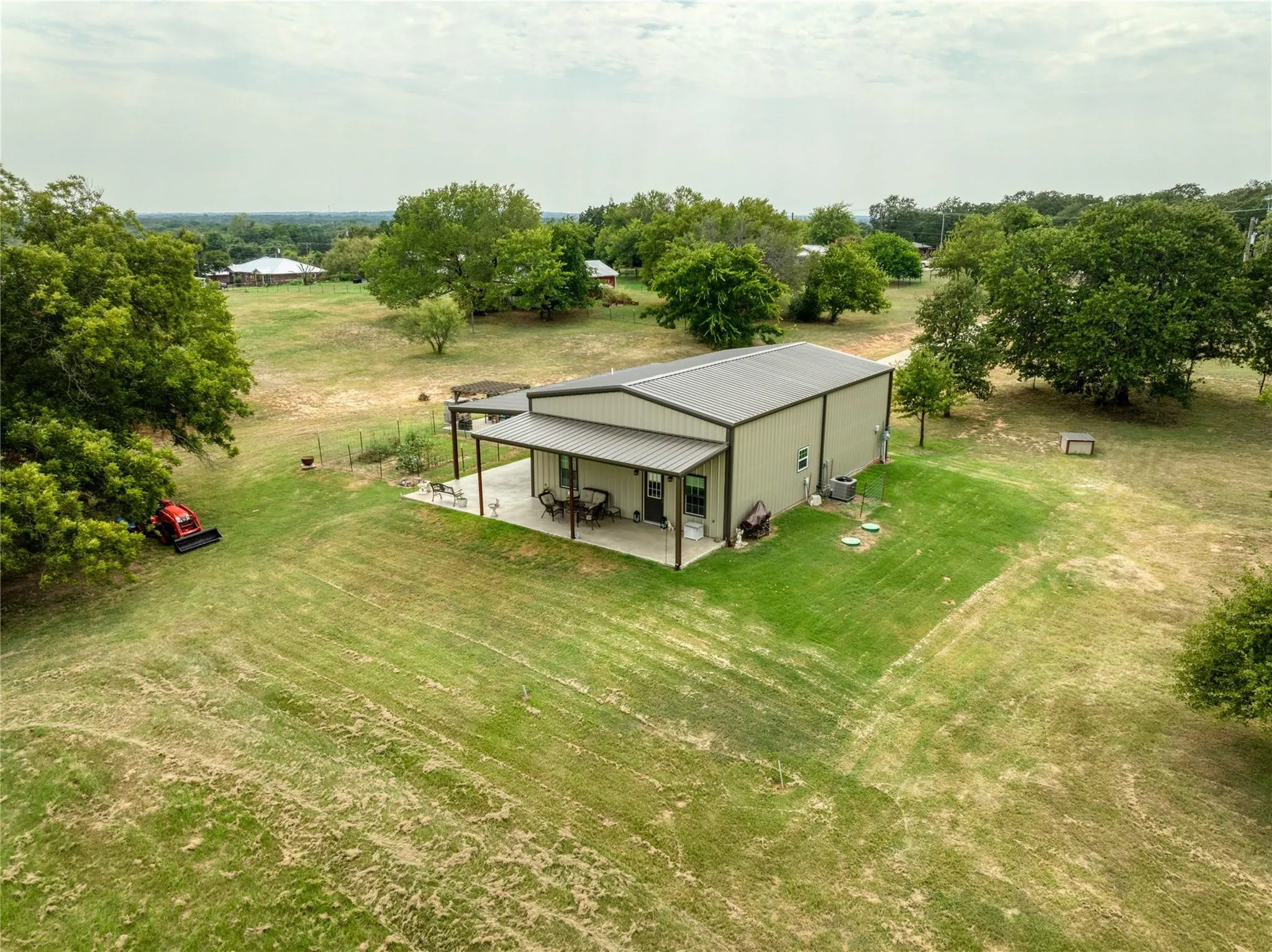 Metal-sided structure featuring a covered concrete patio, an expansive lawn, and mature trees