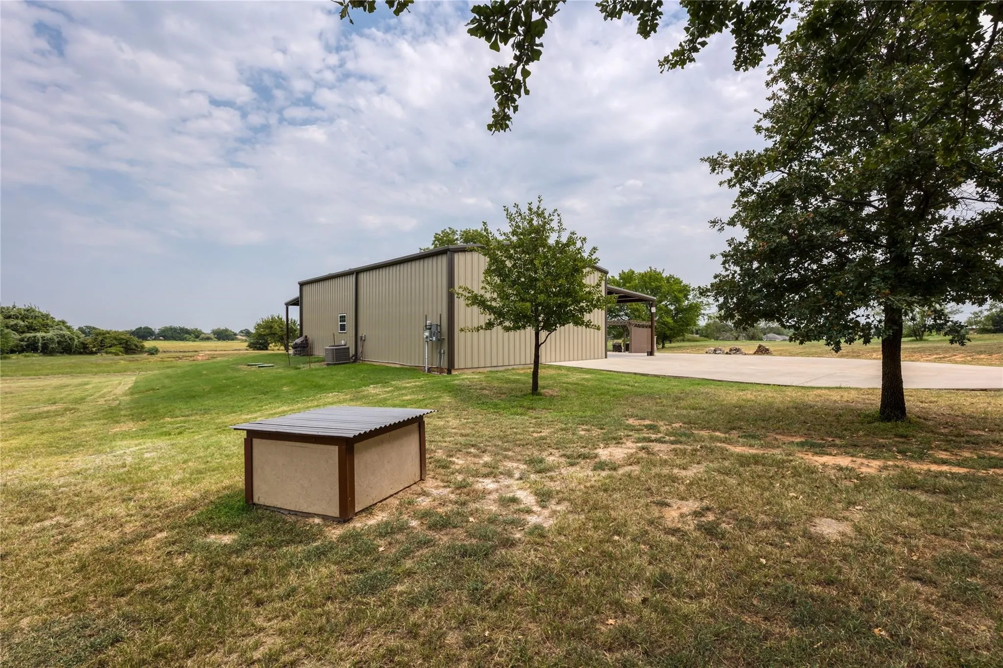 Metal construction building with an attached covered entry, extensive concrete hardscaping, and mature trees