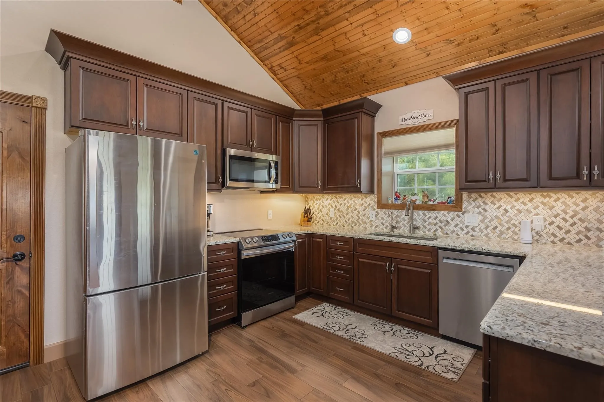 Kitchen featuring wood-finish flooring, dark wood cabinetry, stainless steel appliances, granite countertops, and a mosaic tile backsplash