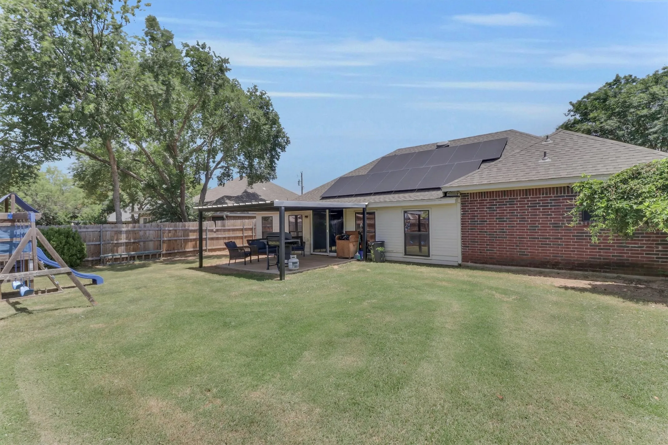 Expansive green lawn with a covered patio, featuring a brick and siding exterior, solar panels, and mature perimeter trees