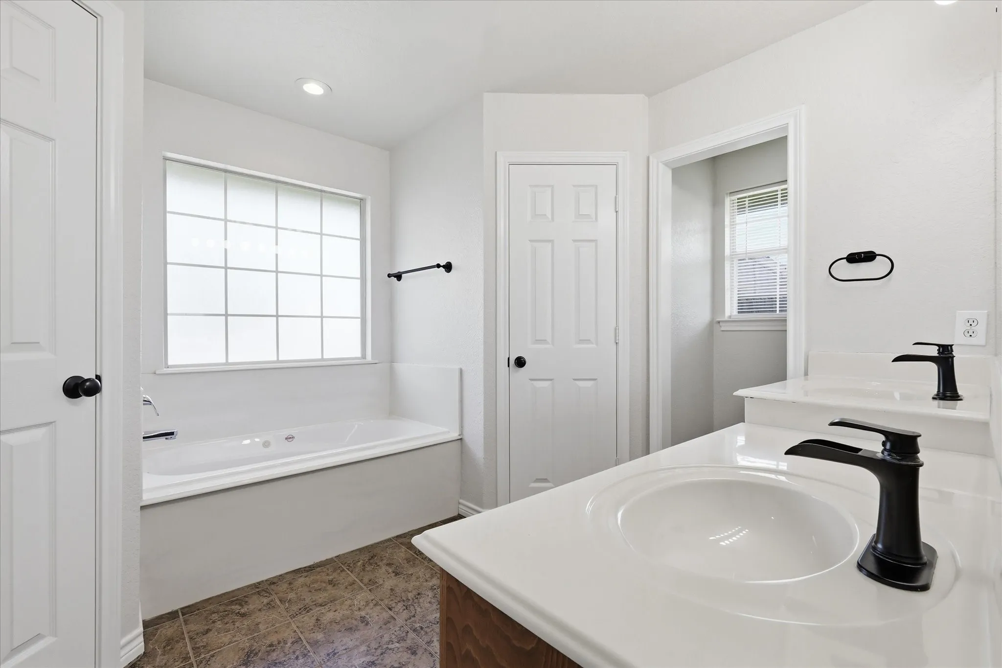 Bathroom featuring a built-in jetted tub under a large window, dual vanities with integrated sinks and matte black fixtures, white panel doors, and tile flooring