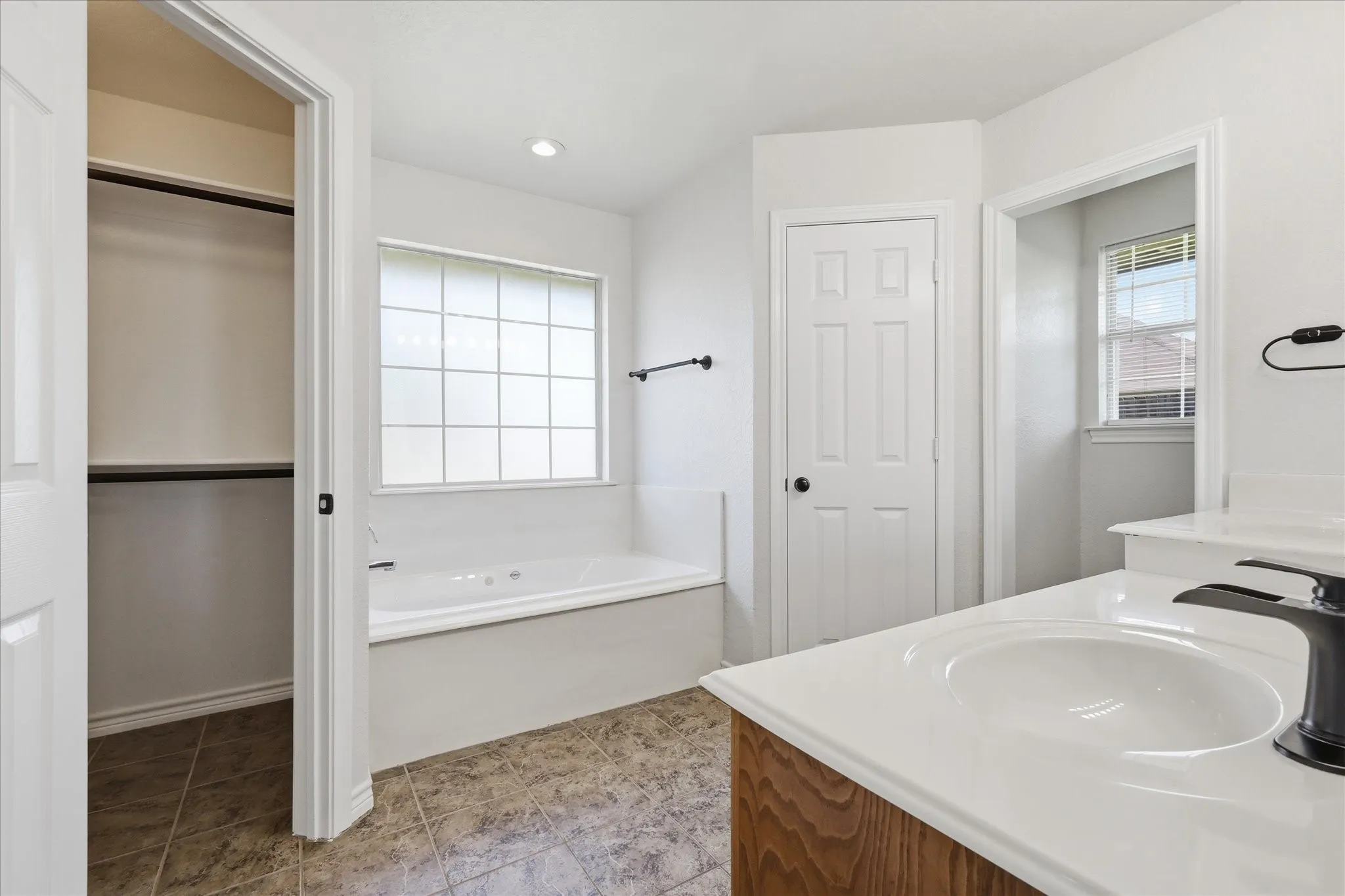 Bathroom featuring a jetted soaking tub with privacy window, dual vanity with undermount sinks and dark hardware, and tile flooring