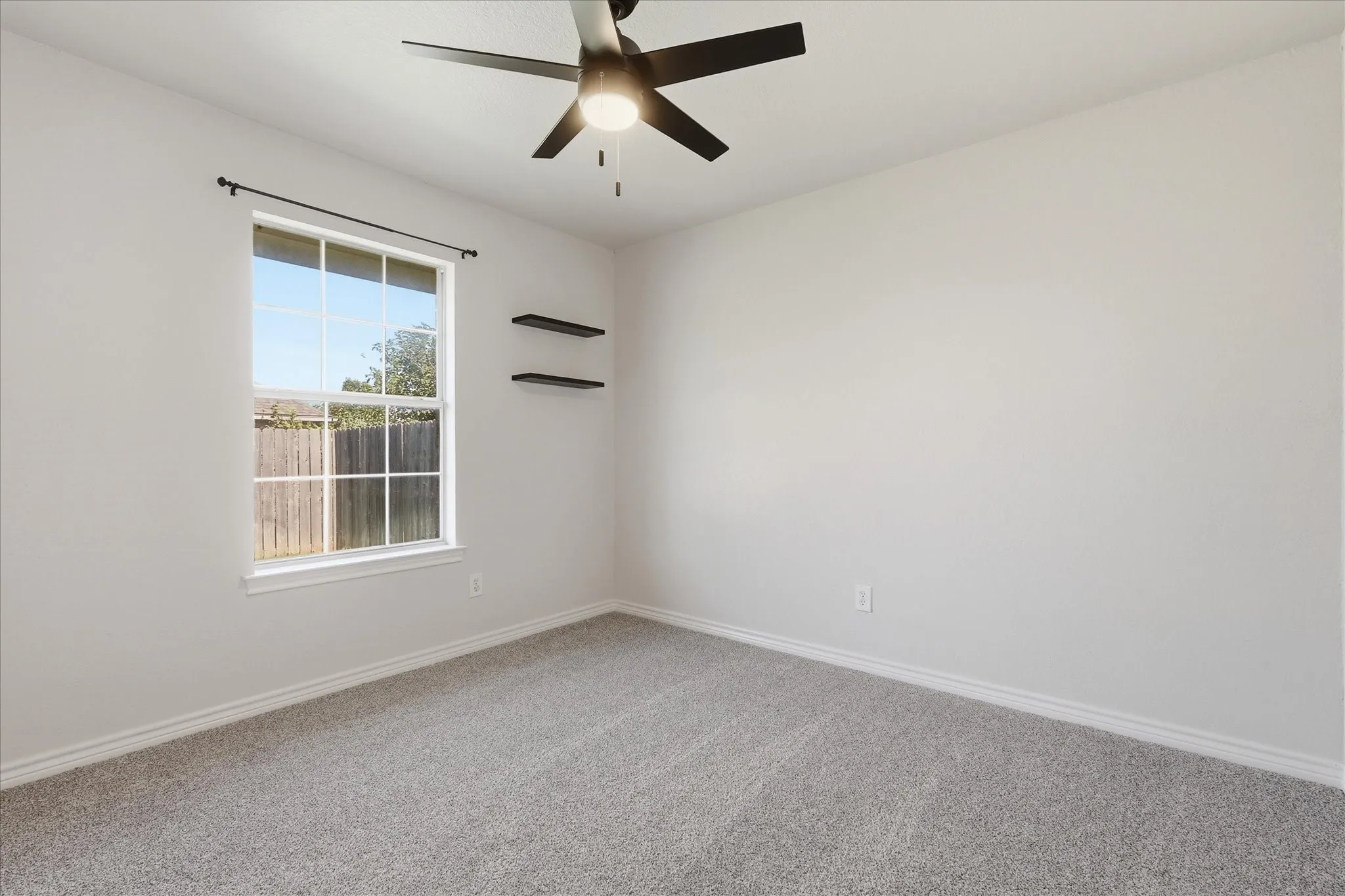 Neutral-toned interior with light gray walls, plush gray carpet, a ceiling fan with integrated lighting, a multi-pane window, and two wall-mounted floating shelves