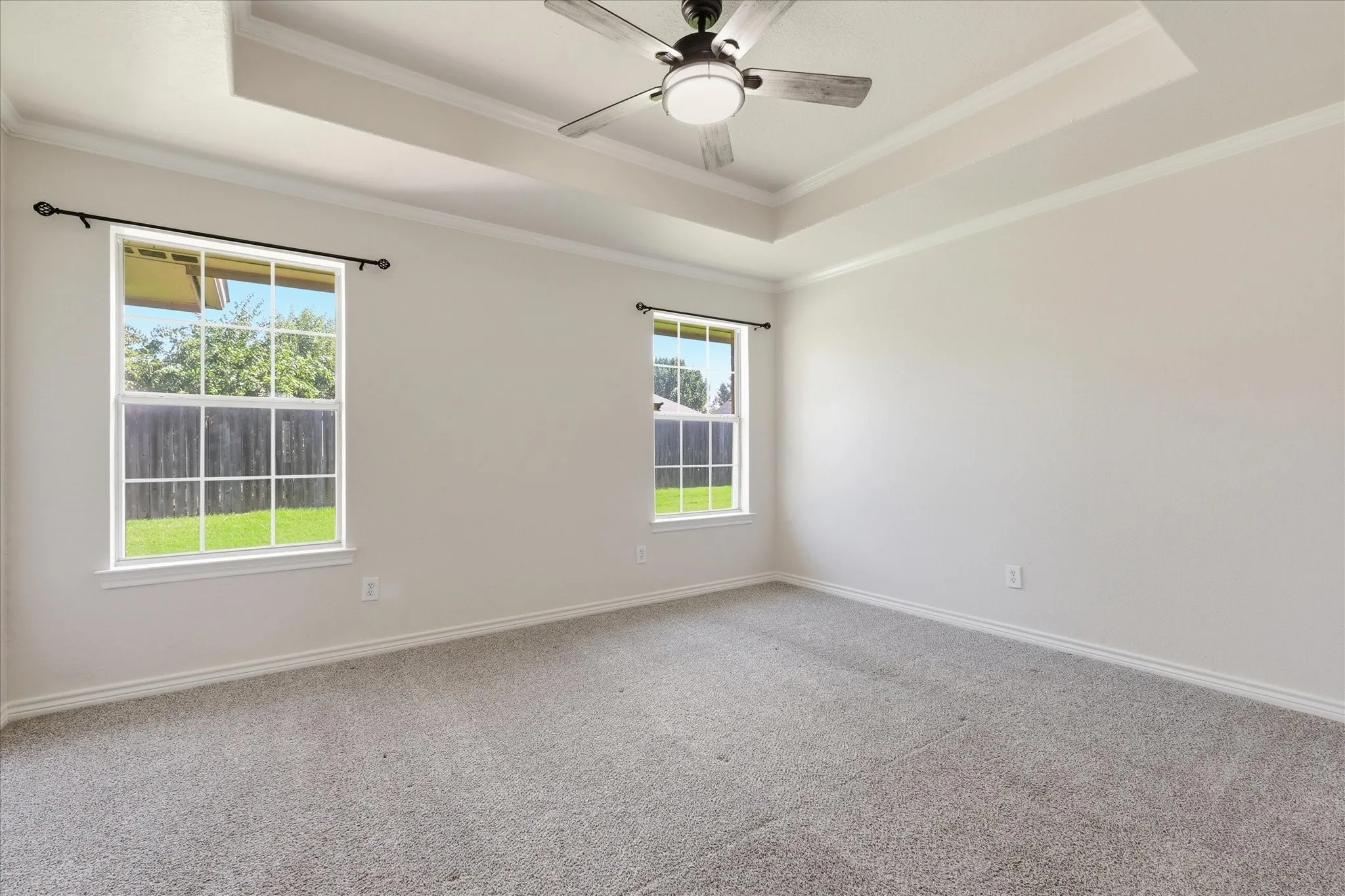 Spacious room featuring a tray ceiling with a ceiling fan, two large windows, light gray carpet flooring, and white baseboards