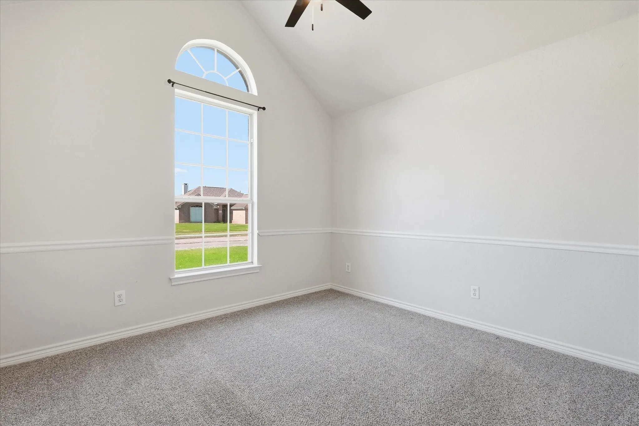 Vaulted ceiling room featuring a large arched top window with grid pattern, neutral gray carpeting, and white chair rail molding