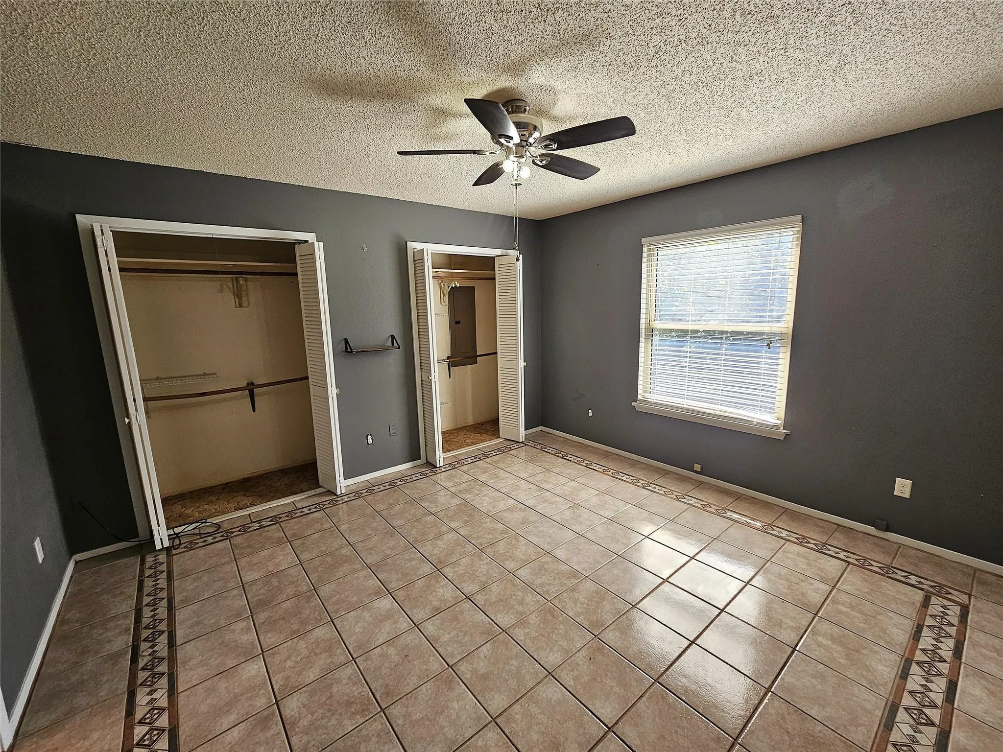 Spacious room featuring neutral tile flooring with decorative inlay, two closets with bi-fold doors, a single window with blinds, and a ceiling fan with integrated lighting