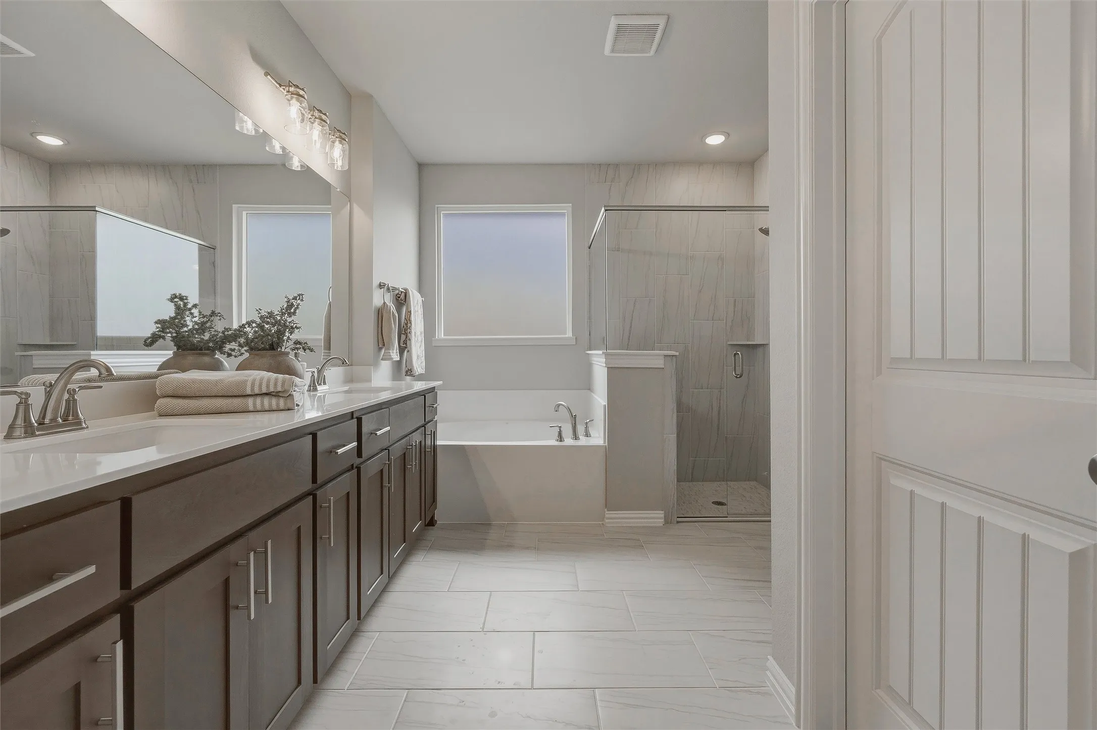 Dual vanity with dark wood-finish cabinetry, a freestanding soaking tub, a glass-enclosed shower, and light-toned tile flooring