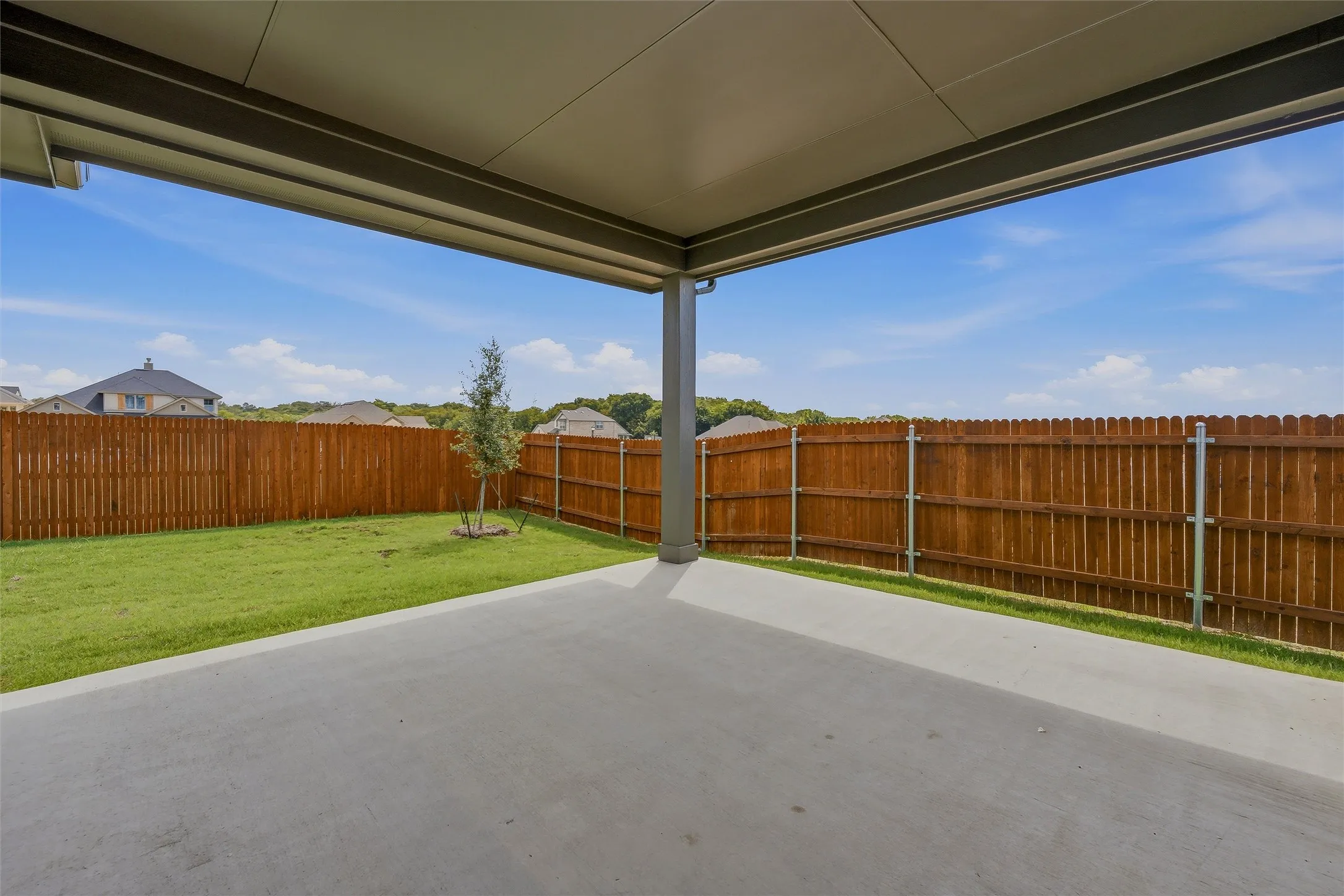 Covered patio with concrete flooring, bordered by a wooden privacy fence and a manicured lawn