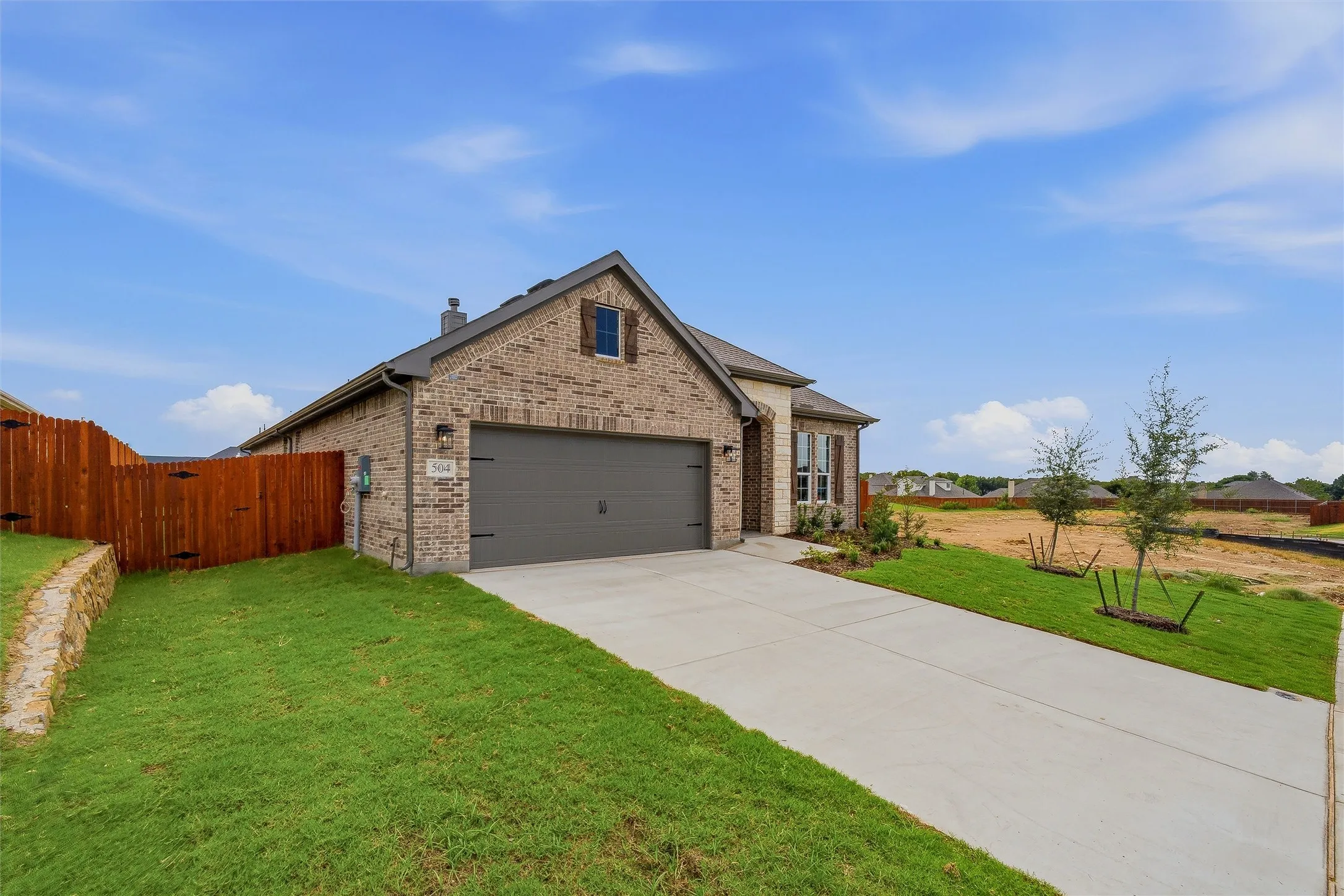 Brick facade exterior with a gabled roofline, an attached garage, a concrete driveway, a wooden privacy fence, and a stone retaining wall
