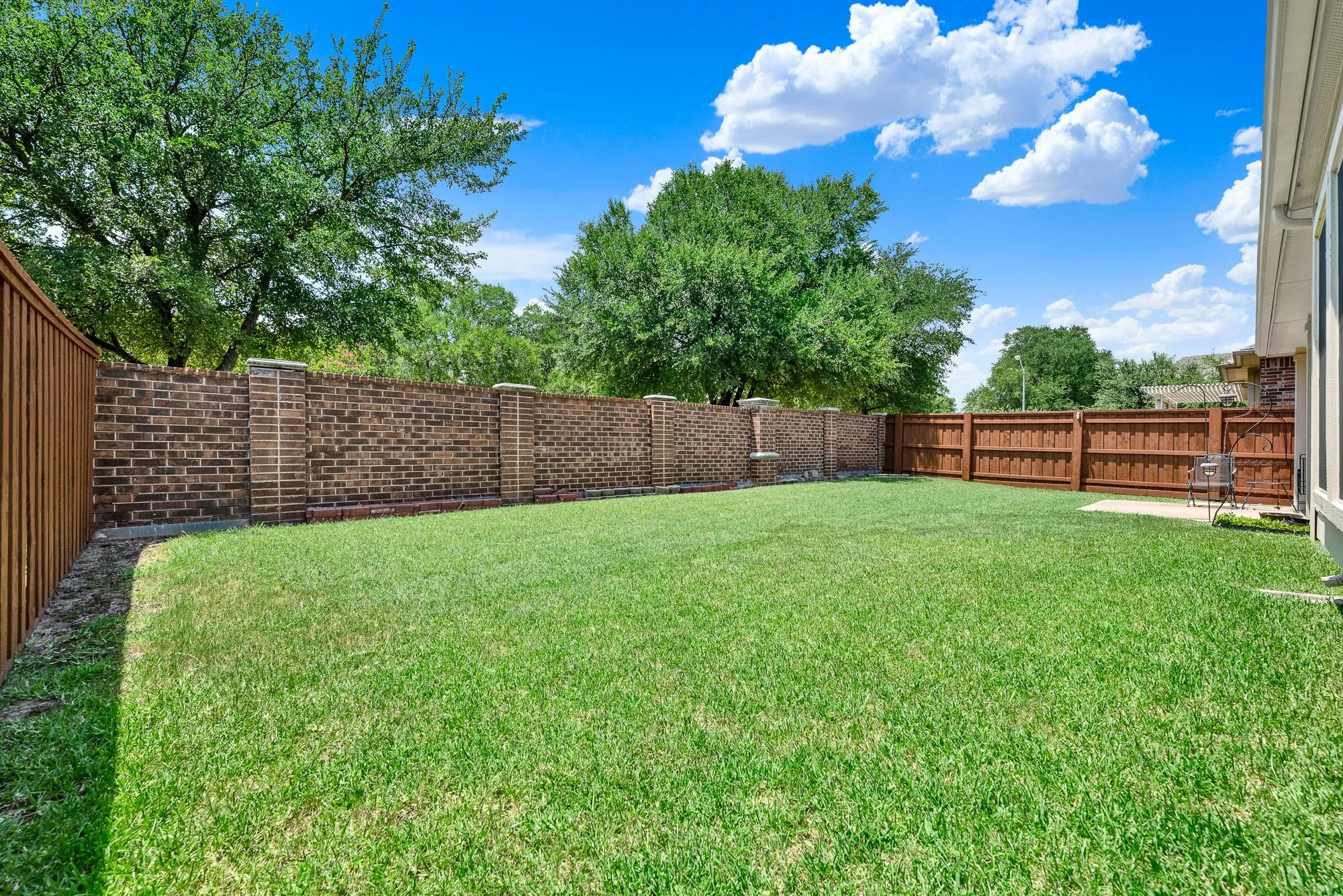 A rear brick privacy wall and mature trees beyond create a peaceful backdrop, enhancing both privacy and the overall outdoor ambiance. The expansive green space is ready for gatherings, games, or quiet evenings outdoors.