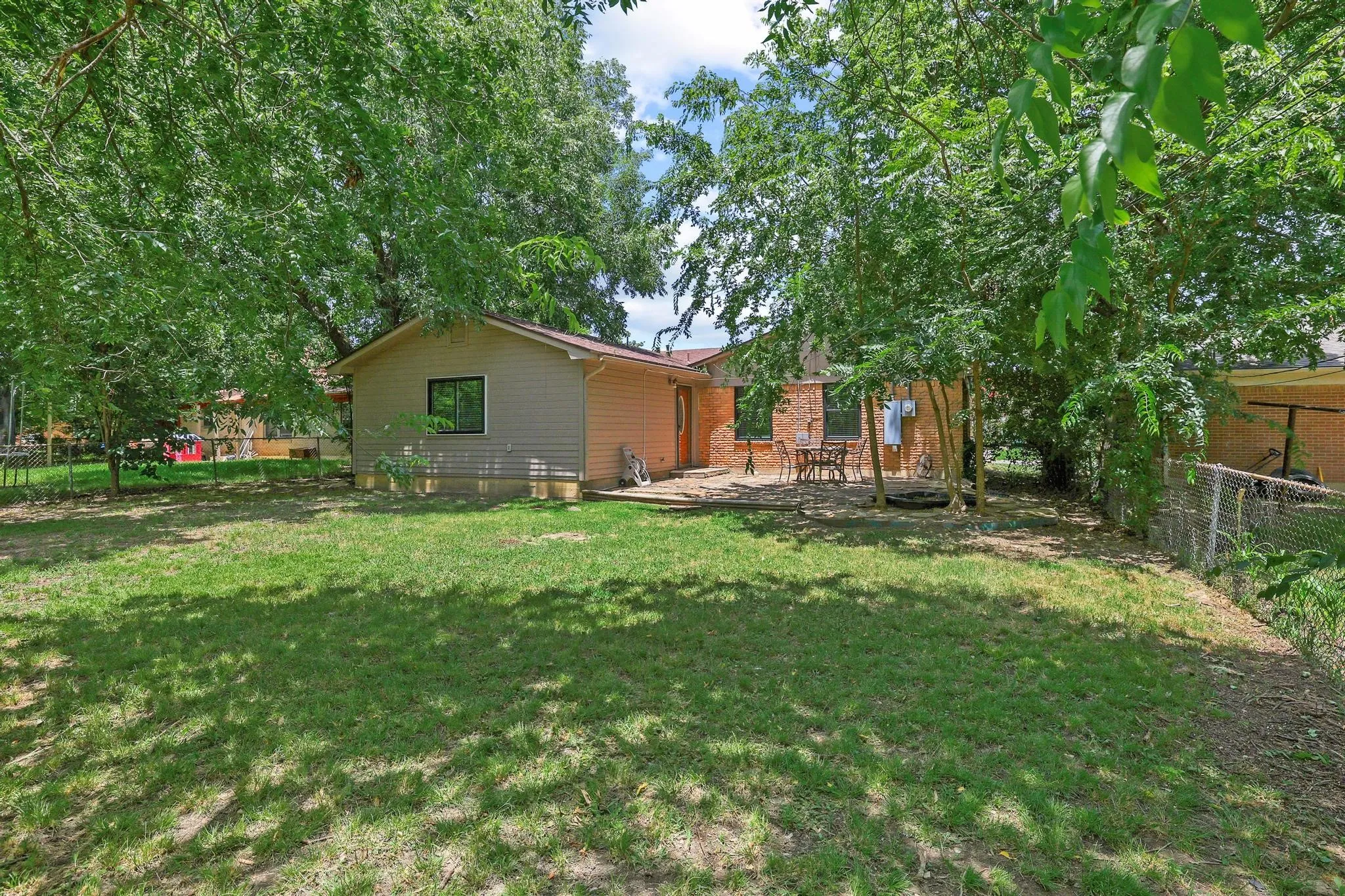 Spacious backyard with mature shade trees, a chain-link fence, and a patio area