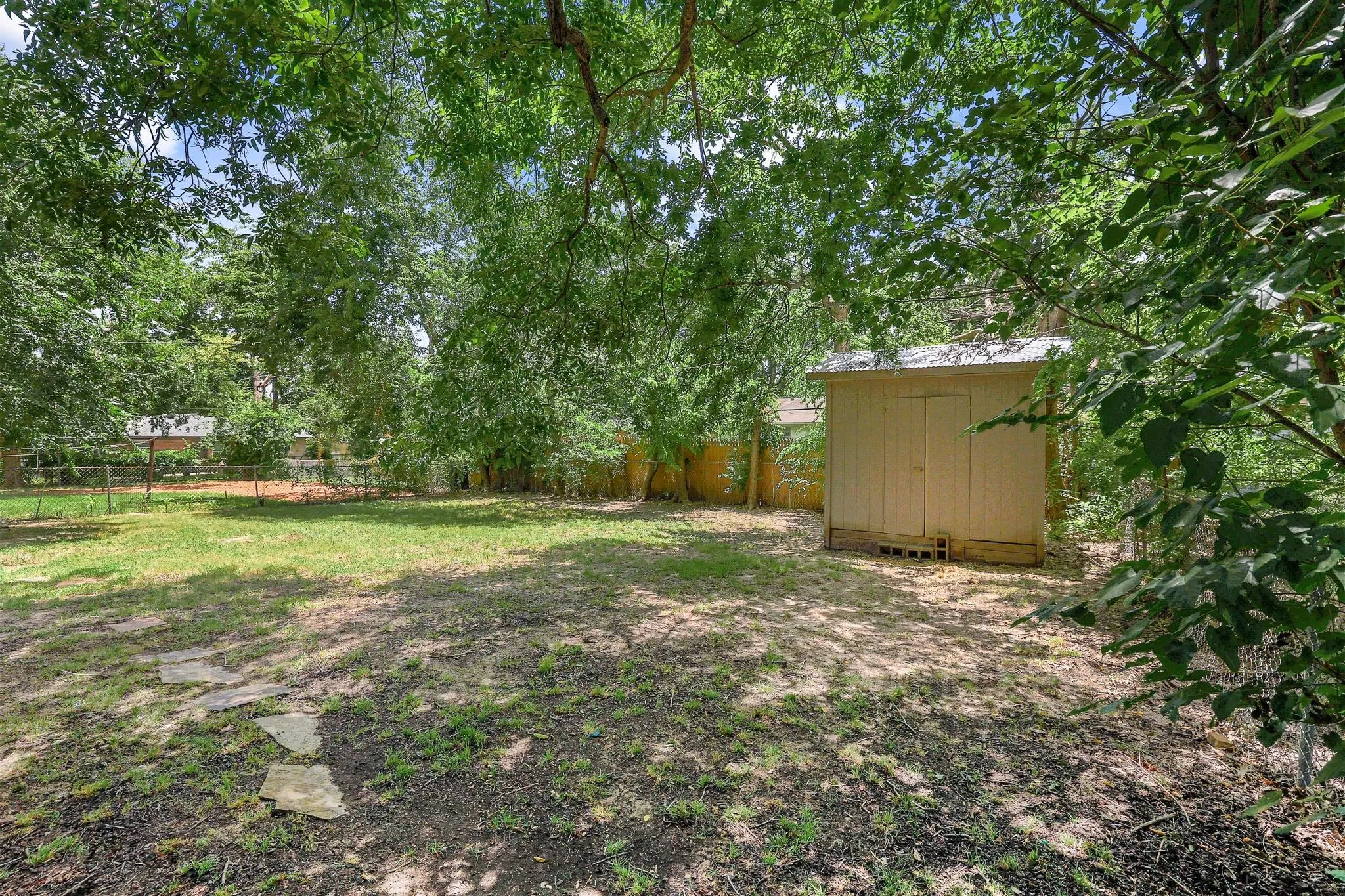 Expansive backyard featuring a storage shed with a metal roof, mature trees providing natural shade, a partial wooden fence, and stone stepping pads