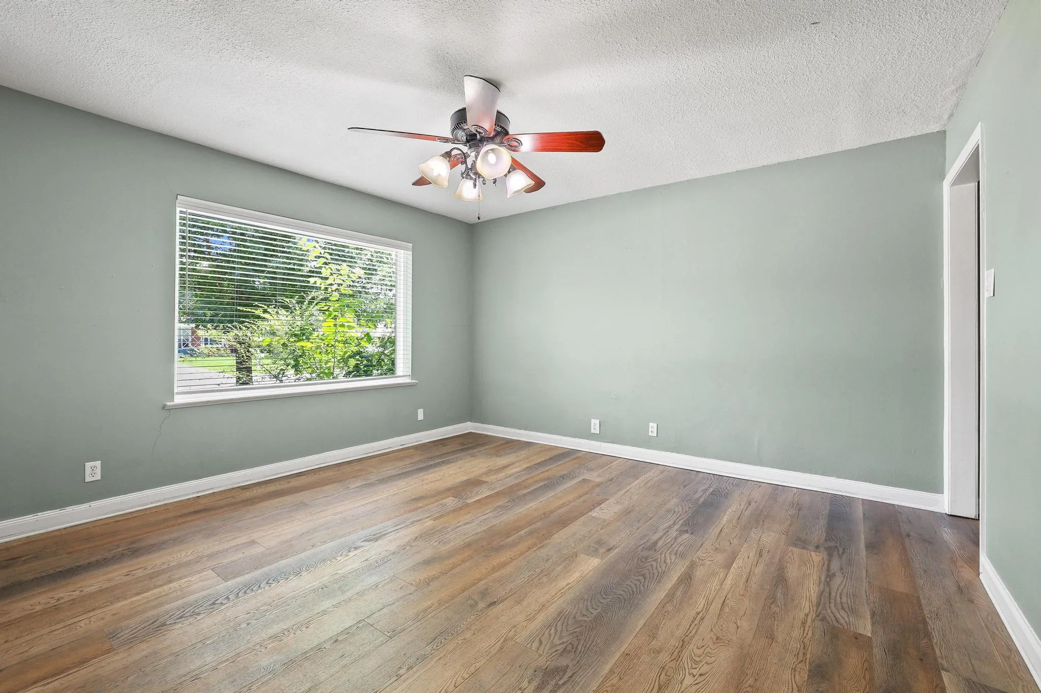 Spacious room featuring wood-finish flooring, a large window with white blinds, a ceiling fan with integrated lighting, and light green wall paint