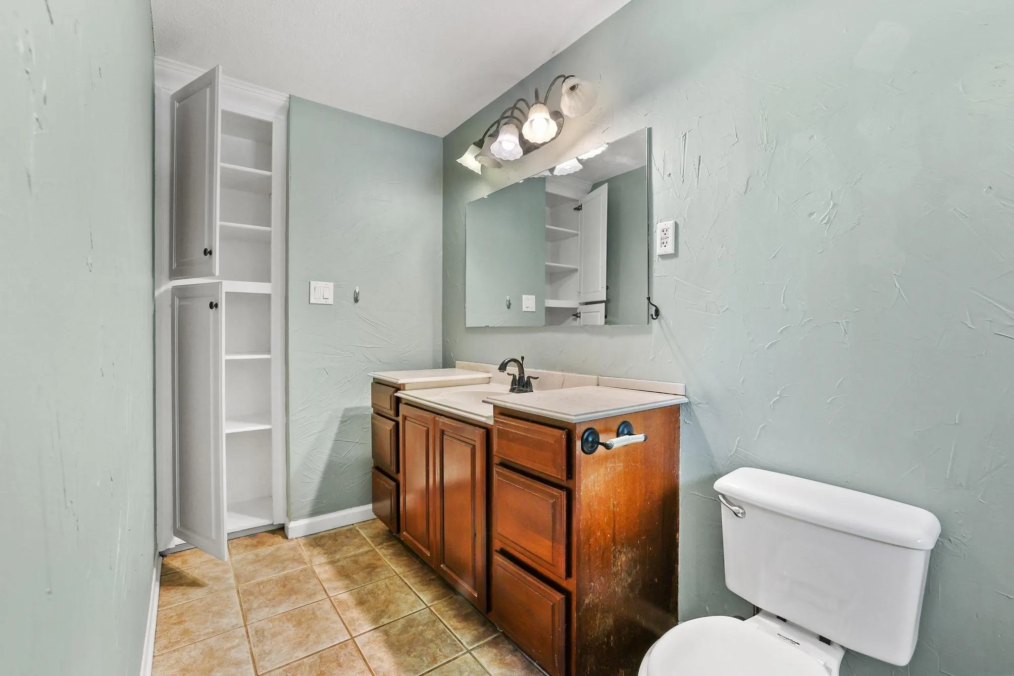 Bathroom featuring a built-in white linen cabinet, single vanity with brown cabinetry and light-toned countertop, wall-mounted mirror, and tile flooring
