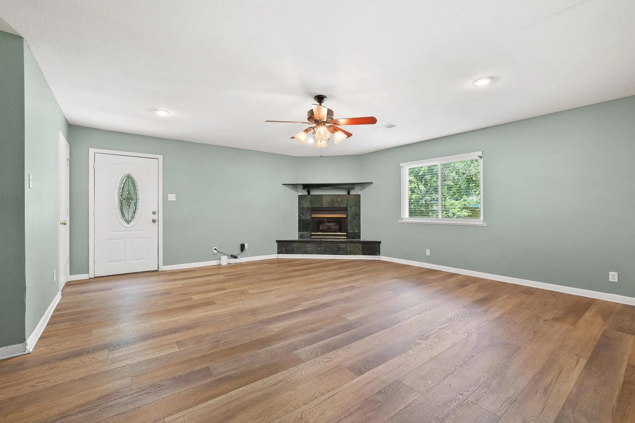 Spacious room featuring wood-finish flooring, a corner fireplace with dark tile surround, a white entry door with decorative glass insert, a ceiling fan, and recessed lighting
