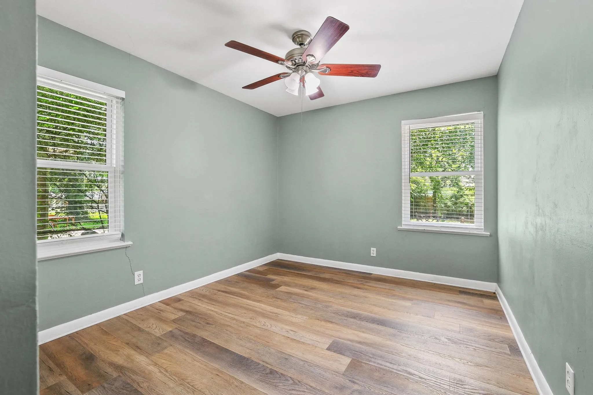 Spacious room featuring wood-finish flooring, two large windows with blinds, a ceiling fan with light fixture, and light green wall tones