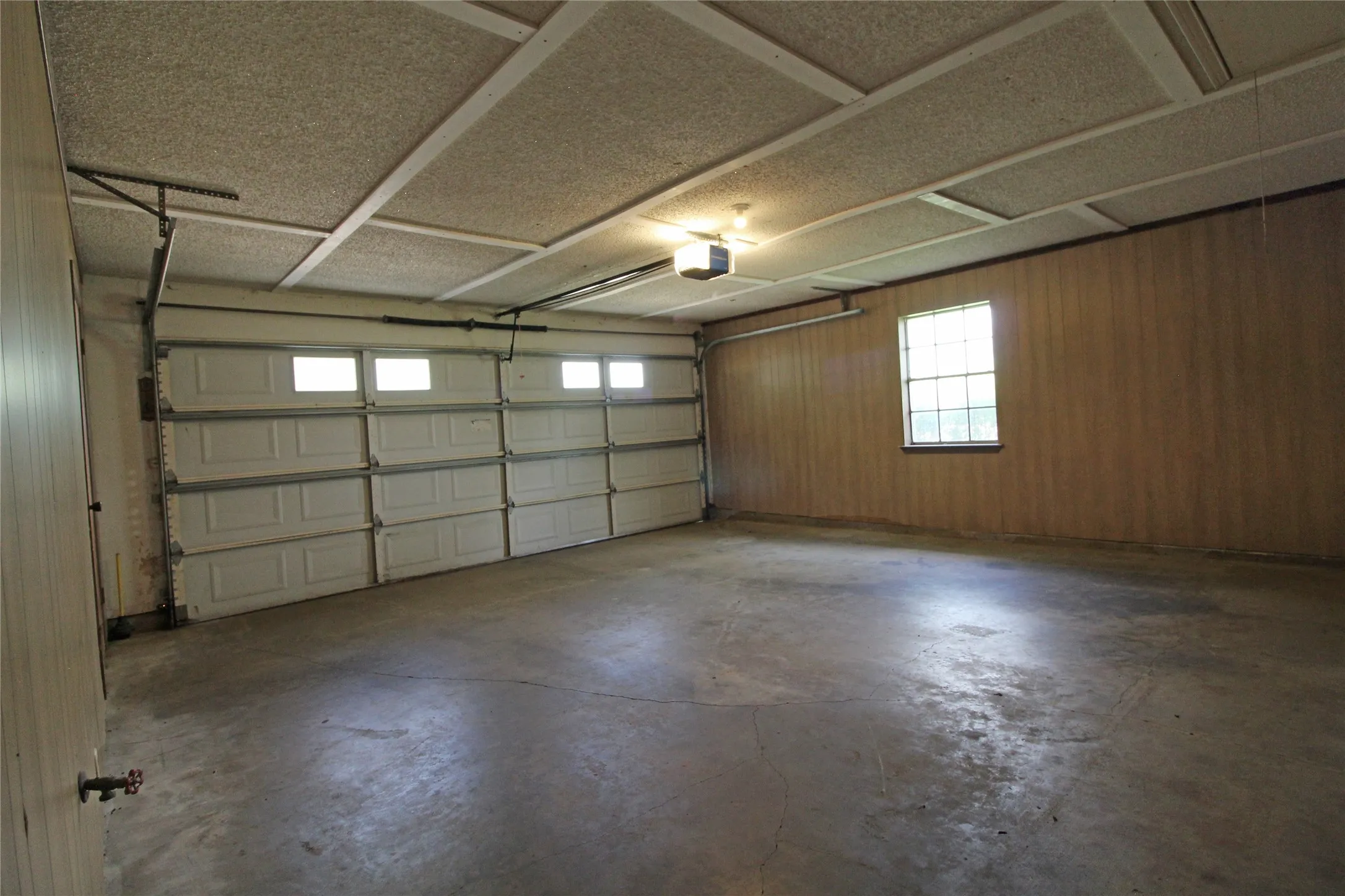 Spacious garage featuring a concrete floor, two white paneled garage doors with upper windows, wood-paneled walls, a window with grilles, and a drop ceiling with acoustic panels