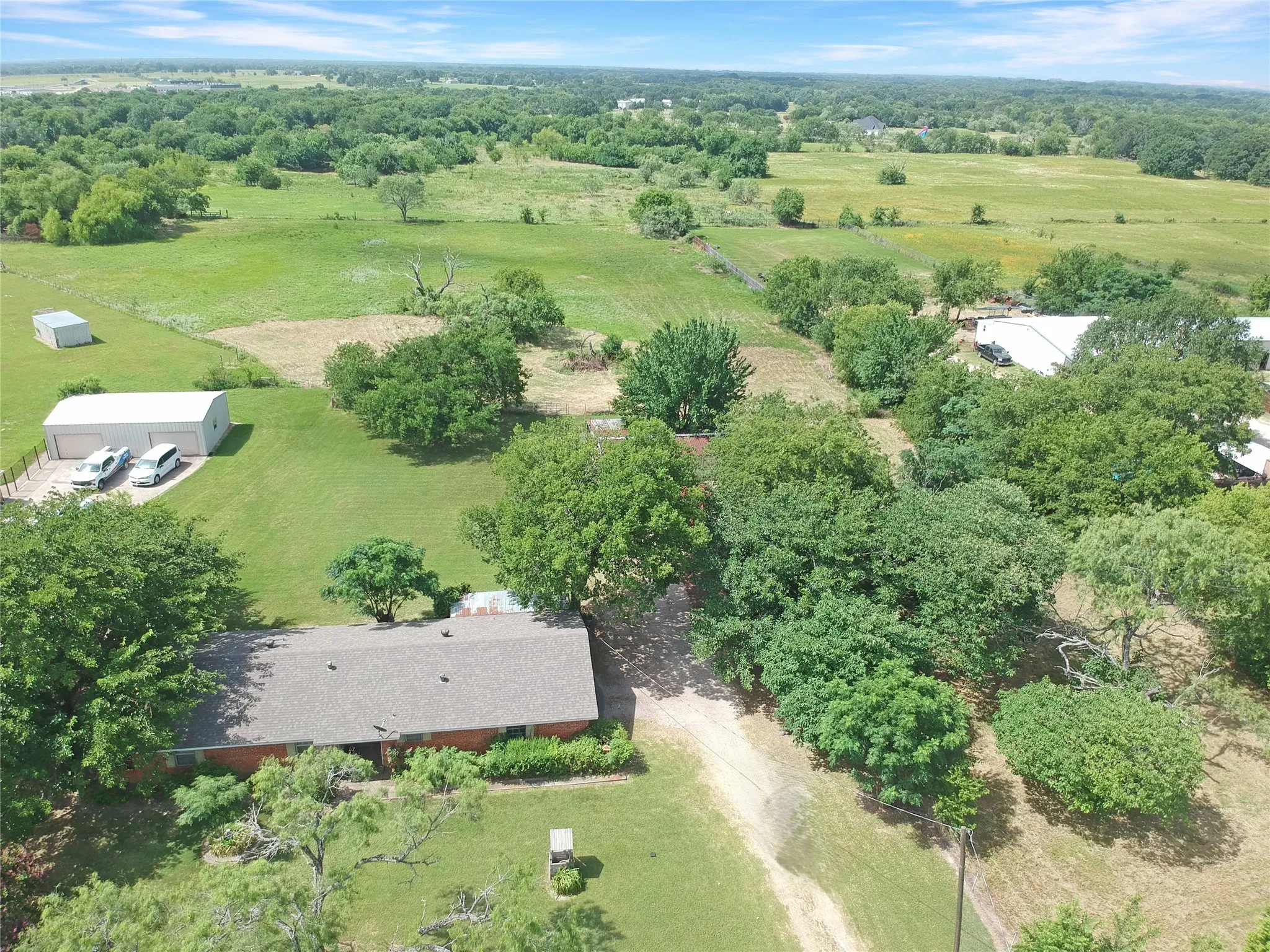 Sprawling property featuring a ranch-style home with a gray shingle roof, surrounded by mature trees and expansive green fields