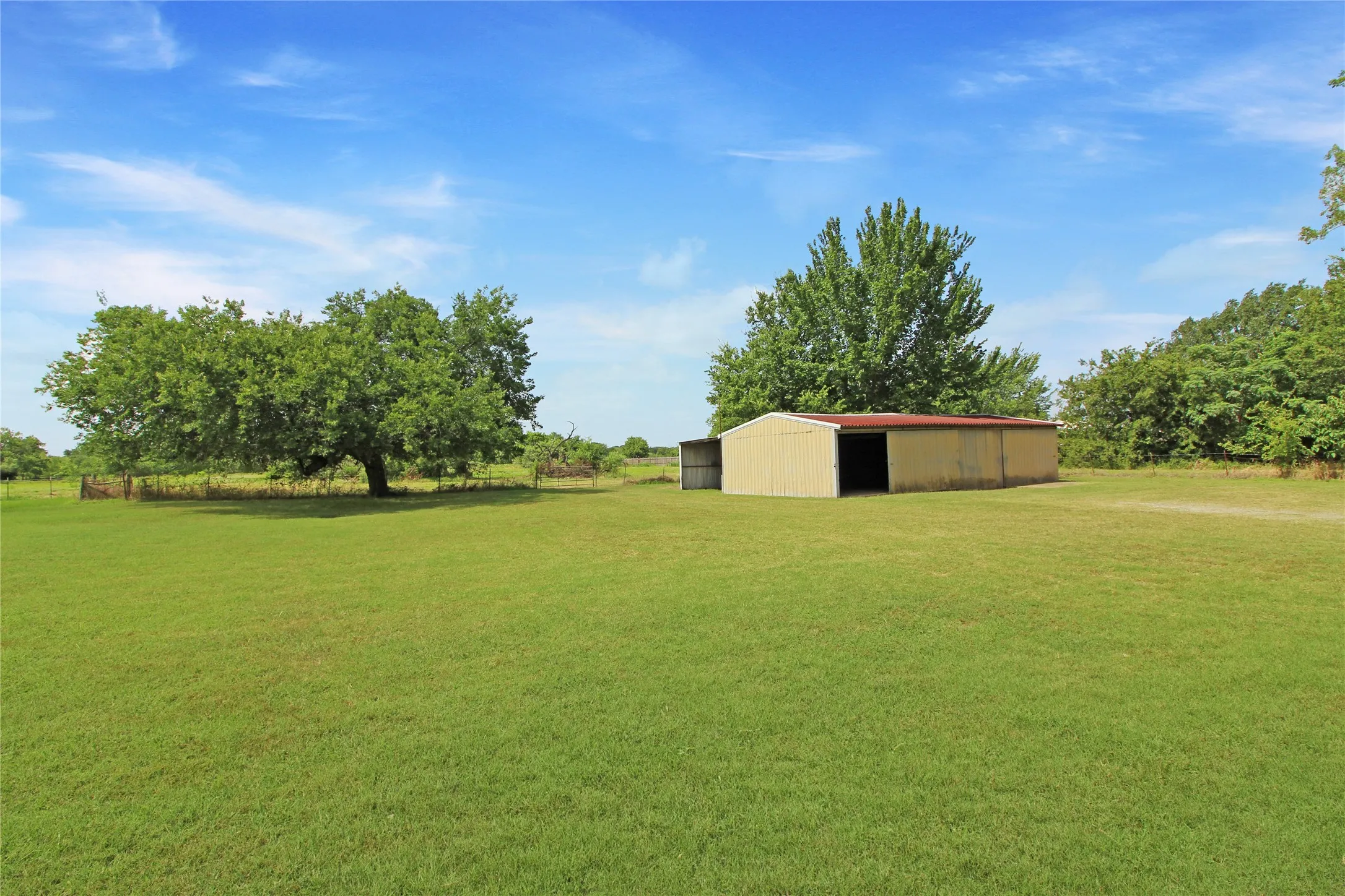 Expansive green lawn featuring a metal outbuilding with open bays, mature trees, and perimeter fencing