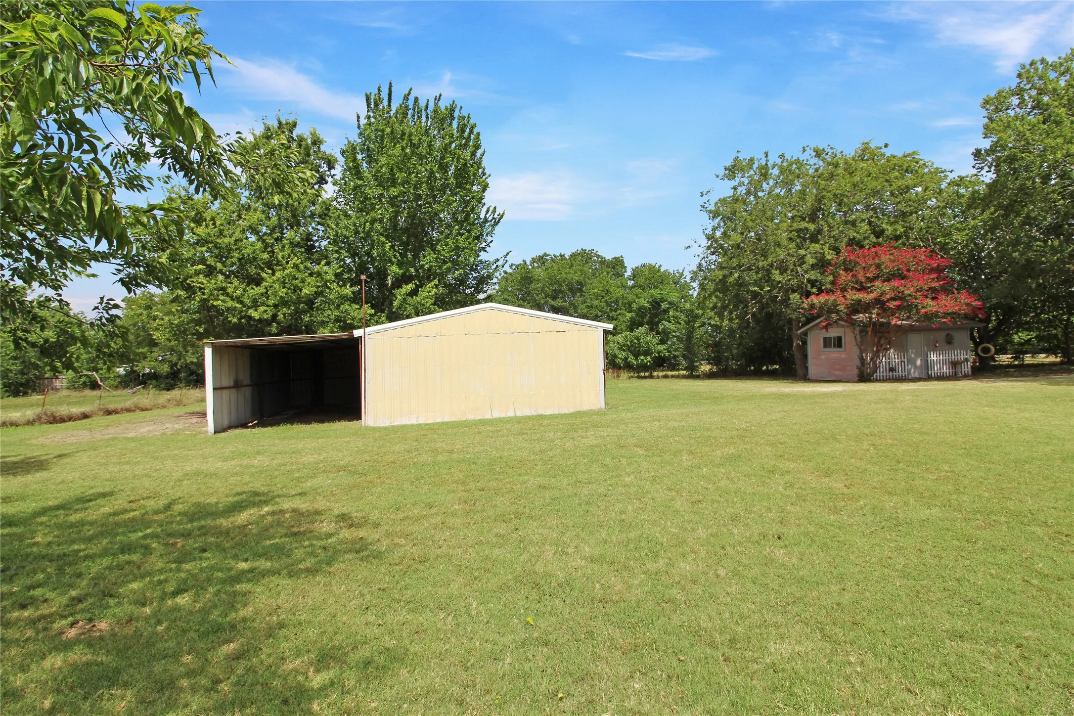 Expansive grassy yard featuring a large open-sided metal utility building and a separate storage shed with a vibrant red-leafed tree